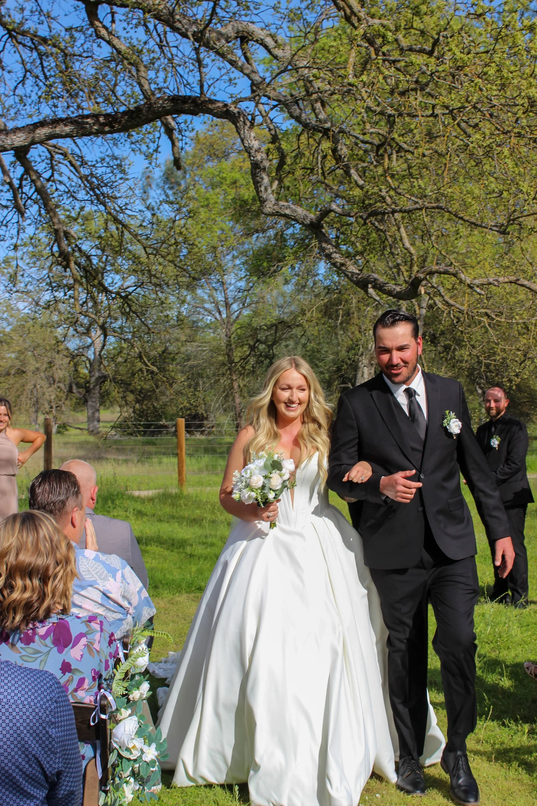 A bride and groom walk arm-in-arm outdoors during their wedding ceremony, smiling. The bride holds a bouquet of white flowers, and guests sit on the grass, watching and smiling. There are large trees and a fence in the background, with a clear blue s