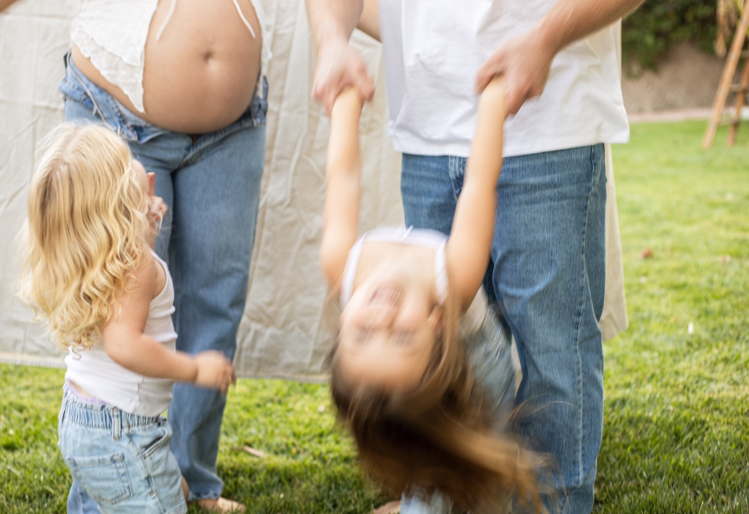 A pregnant woman and a man holding a child's hands while the child dips backward at an outdoor gathering, with a pregnant belly visible and all three in casual clothing on grass.