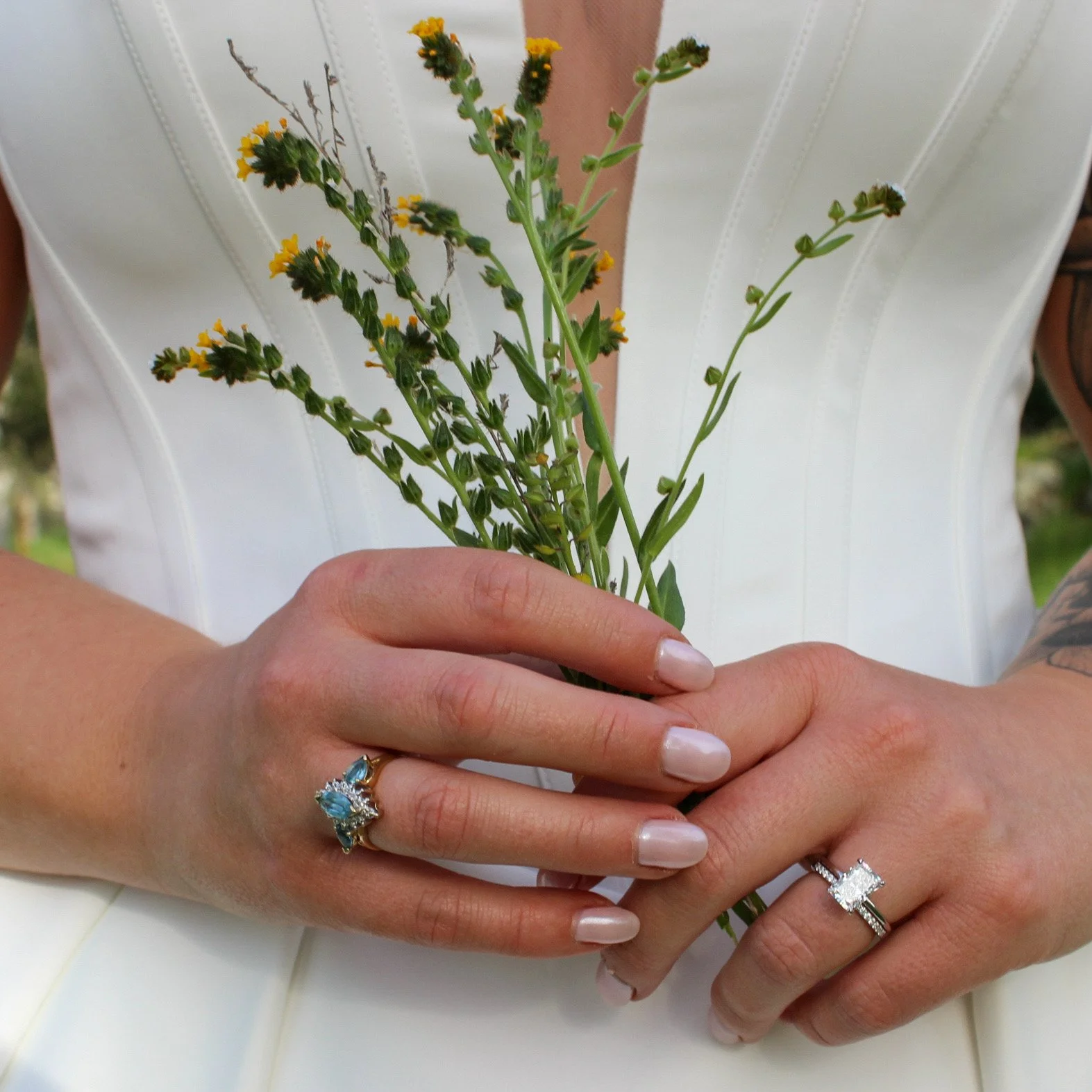 Person holding a small bouquet of wildflowers, wearing a white dress with a deep V neckline, and displaying a large engagement ring on their right hand and a decorative ring on their left hand.
