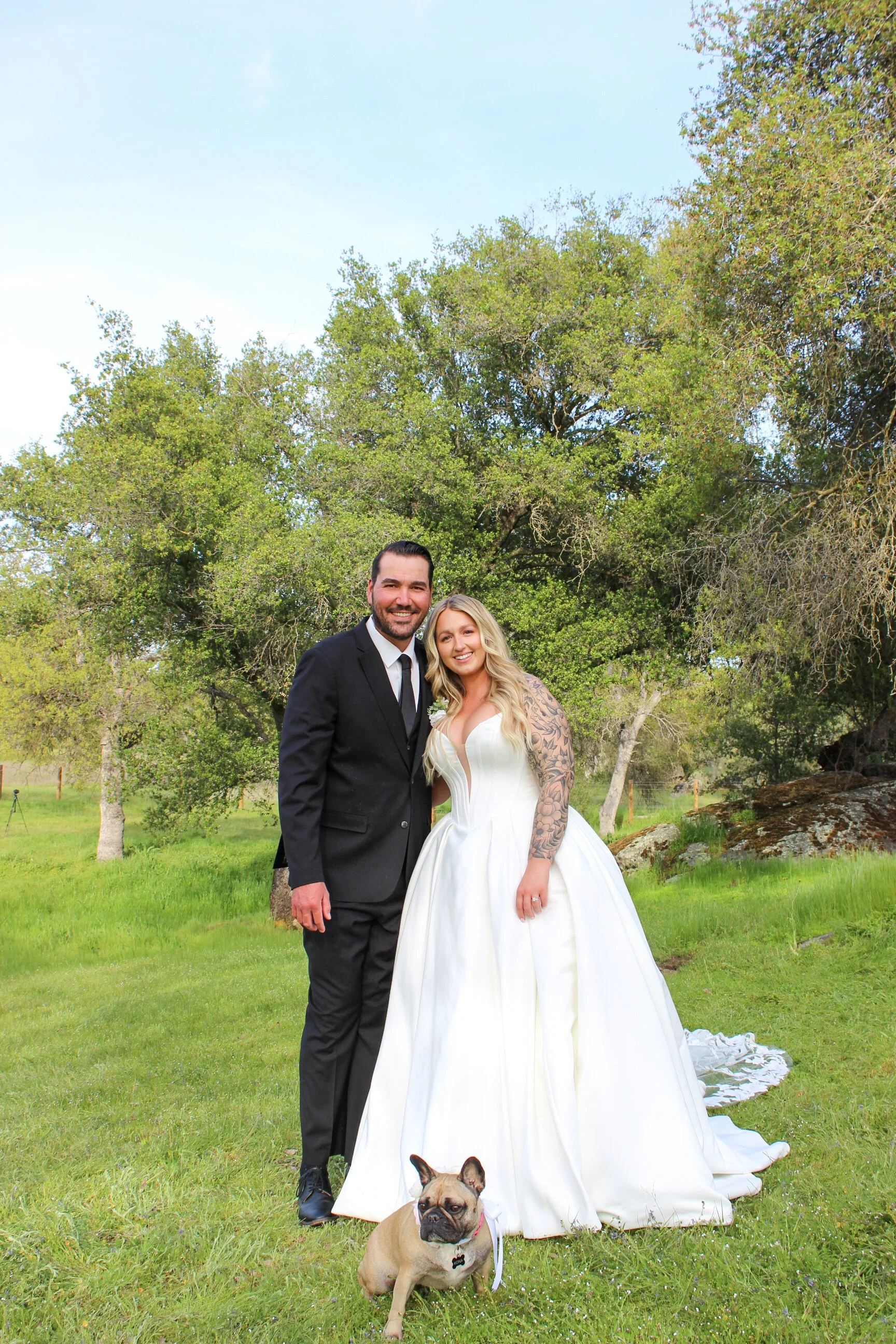 A newly married couple, dressed in wedding attire, standing outdoors on a grassy area with trees in the background. The groom is wearing a black suit and tie, and the bride is wearing a white wedding gown with tattoos on her arm. They are smiling and