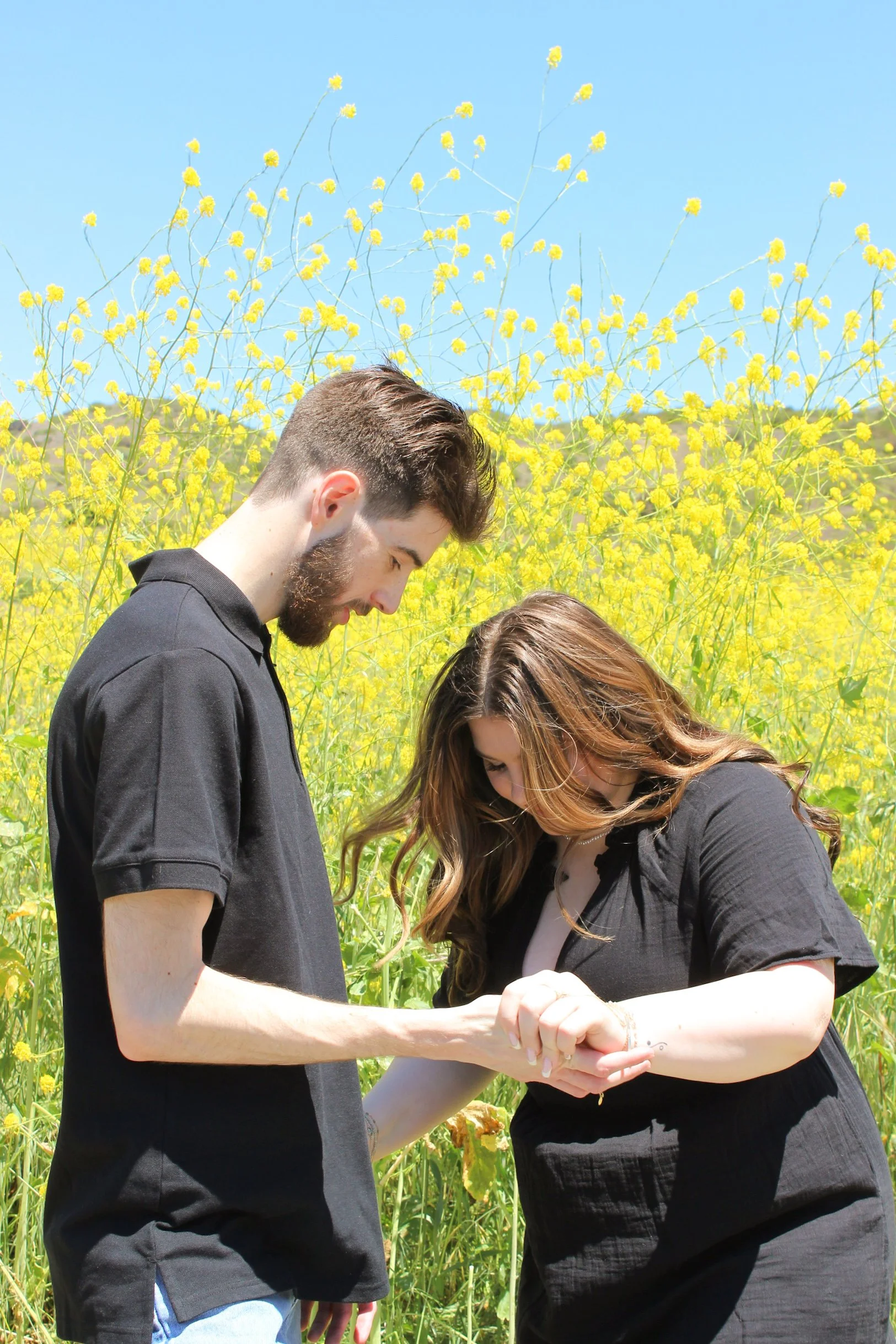 A young man and woman standing close together outdoors, looking at each other and holding hands, in front of a field of yellow flowers with a clear blue sky above.