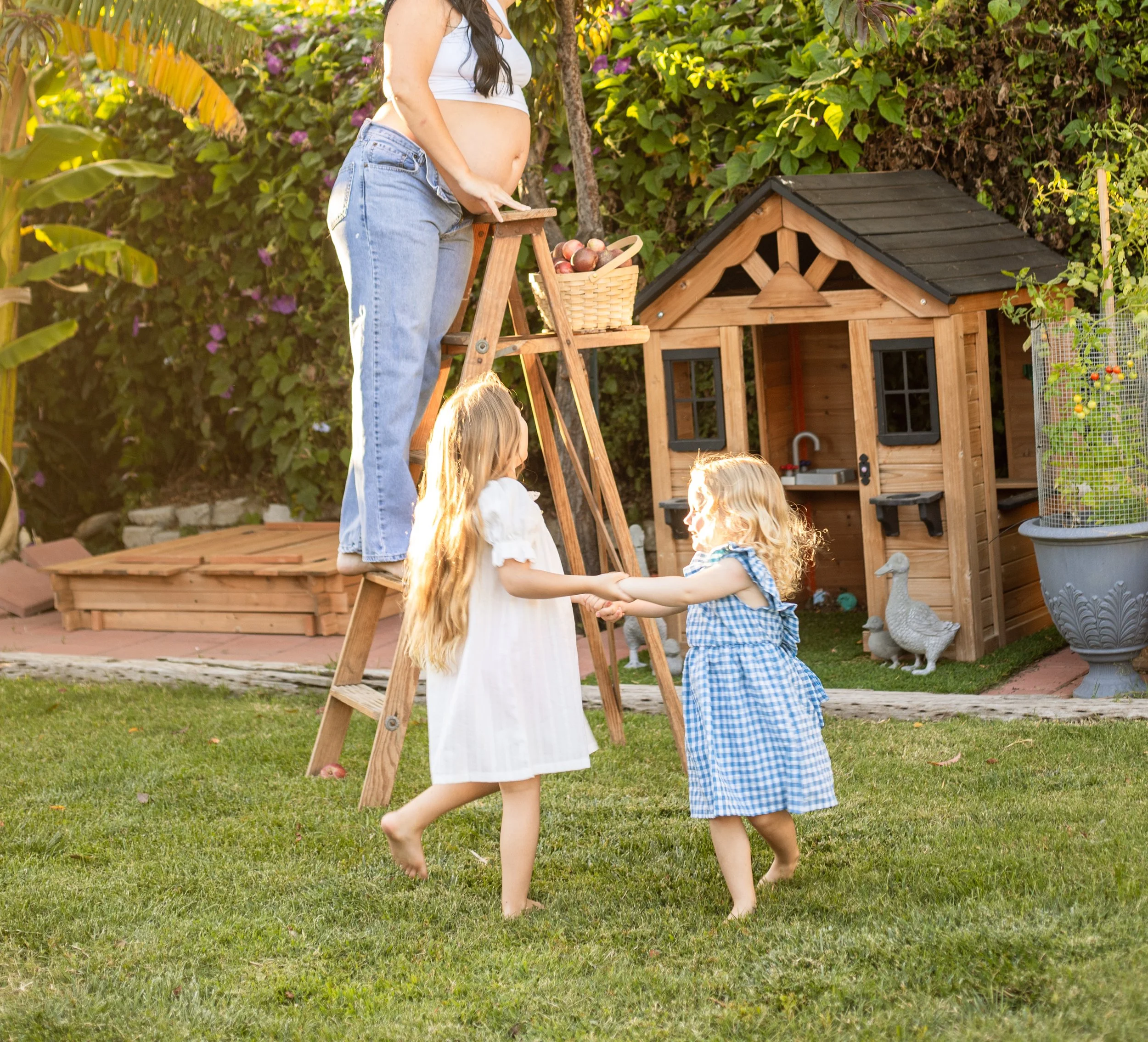 Two young girls are dancing and holding hands in a garden, while an adult woman, standing on a ladder, picks apples from a tree. The background features a wooden playhouse, a basket of apples, and decorative ducks.