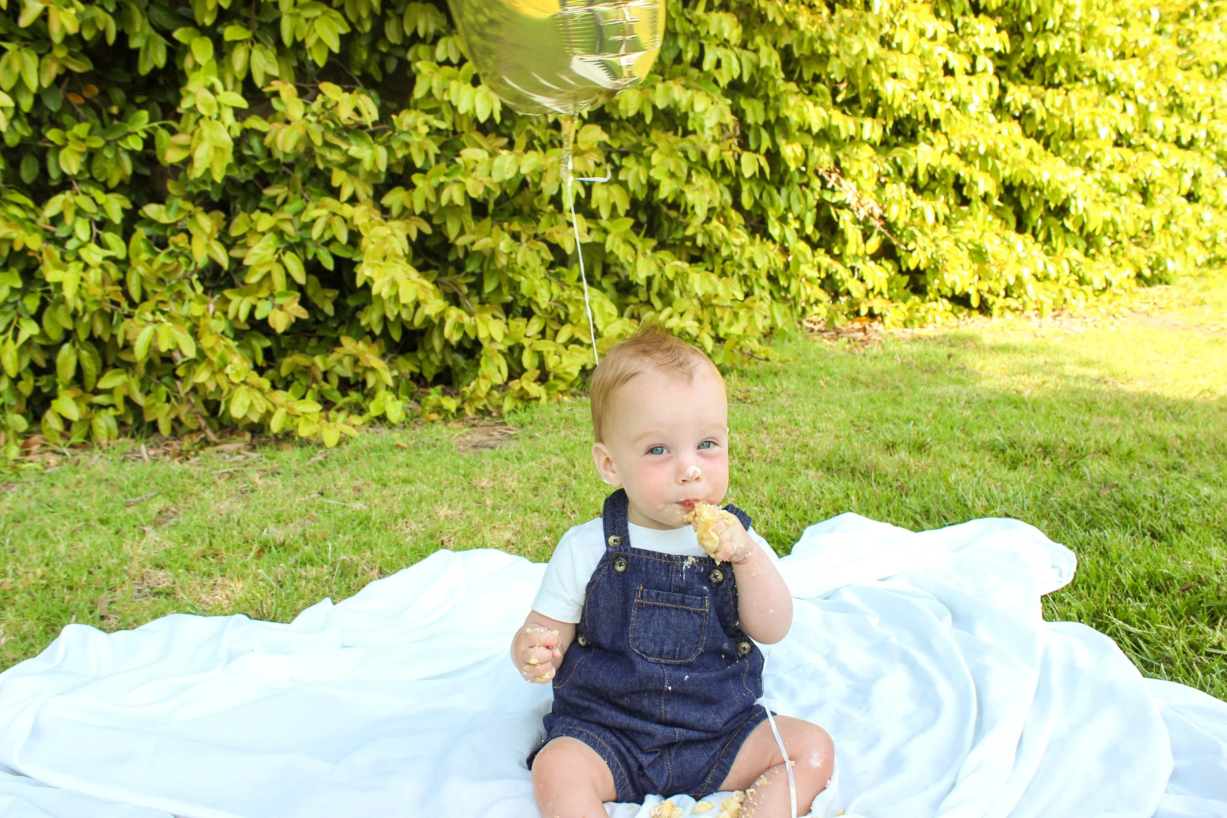 A young child with light hair and blue eyes sitting on a white blanket on the grass, eating cake with some frosting on the face, outdoors with green bushes in the background, holding a balloon.