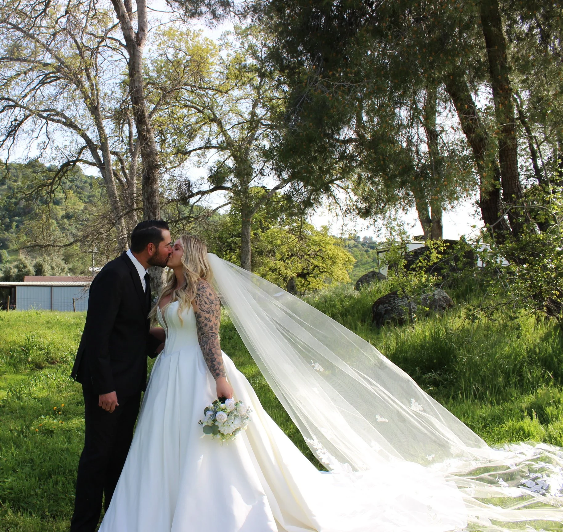 A bride and groom share a kiss outdoors in a green, wooded area on their wedding day.