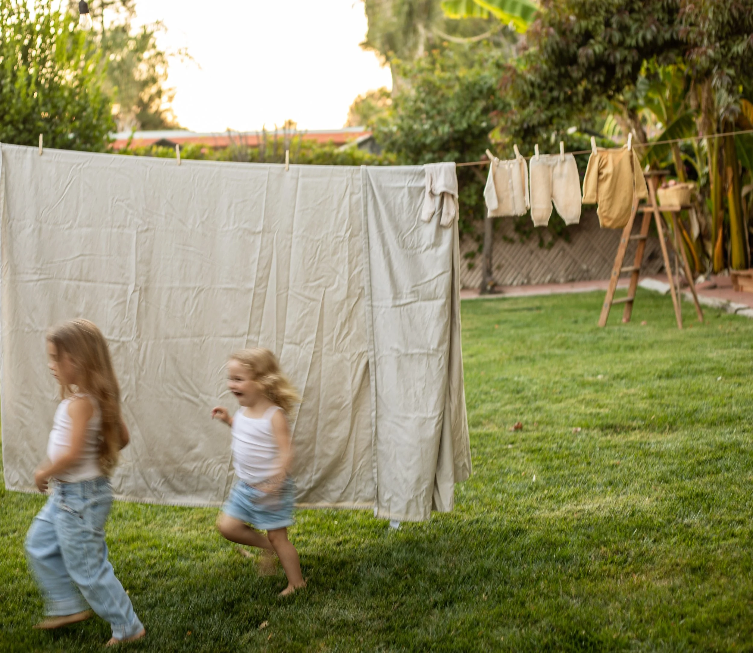 Two young girls running and laughing in a backyard, with a laundry line holding laundry in the background.