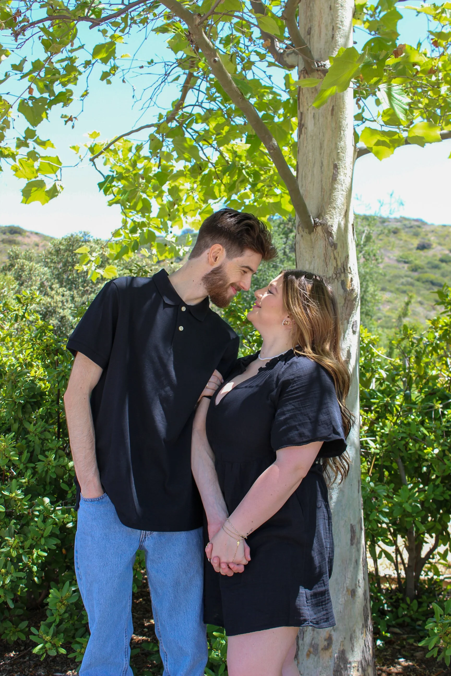 A young couple with fair skin holding hands and leaning against a tree outdoors, smiling and gazing at each other on a sunny day.