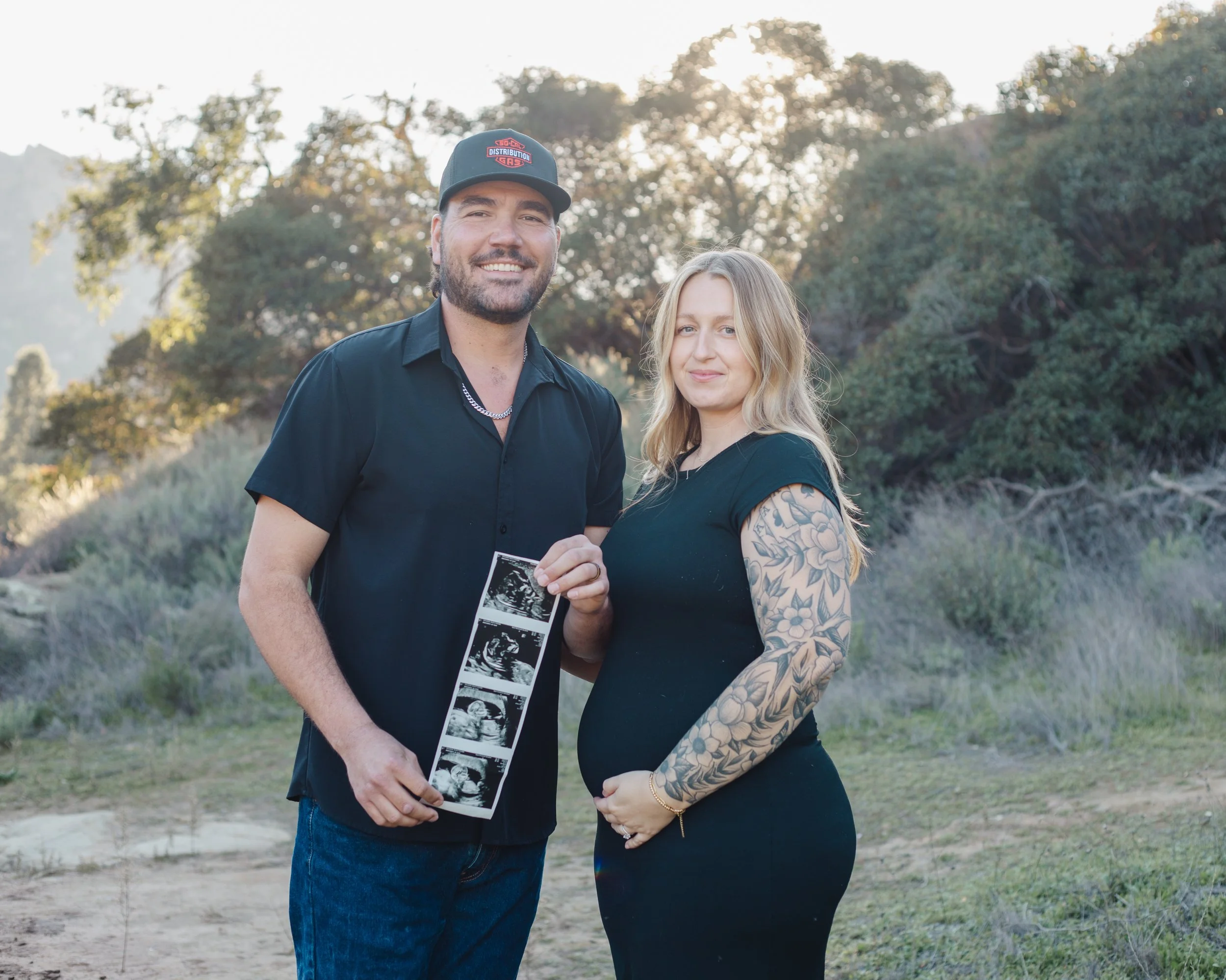 A smiling man holding ultrasound images and a pregnant woman with tattoos on her arm, standing outdoors during sunset.