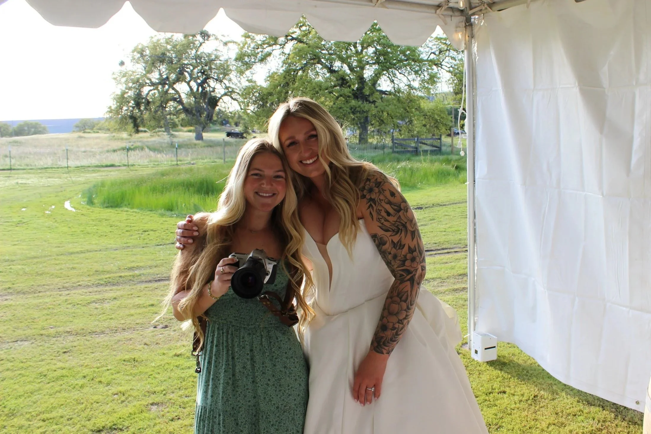 Two women standing together outdoors under a white canopy tent, smiling at the camera. One woman is wearing a white dress with tattoos on her arm, and the other is wearing a green dress and holding a camera. Green fields and trees are visible in the background.