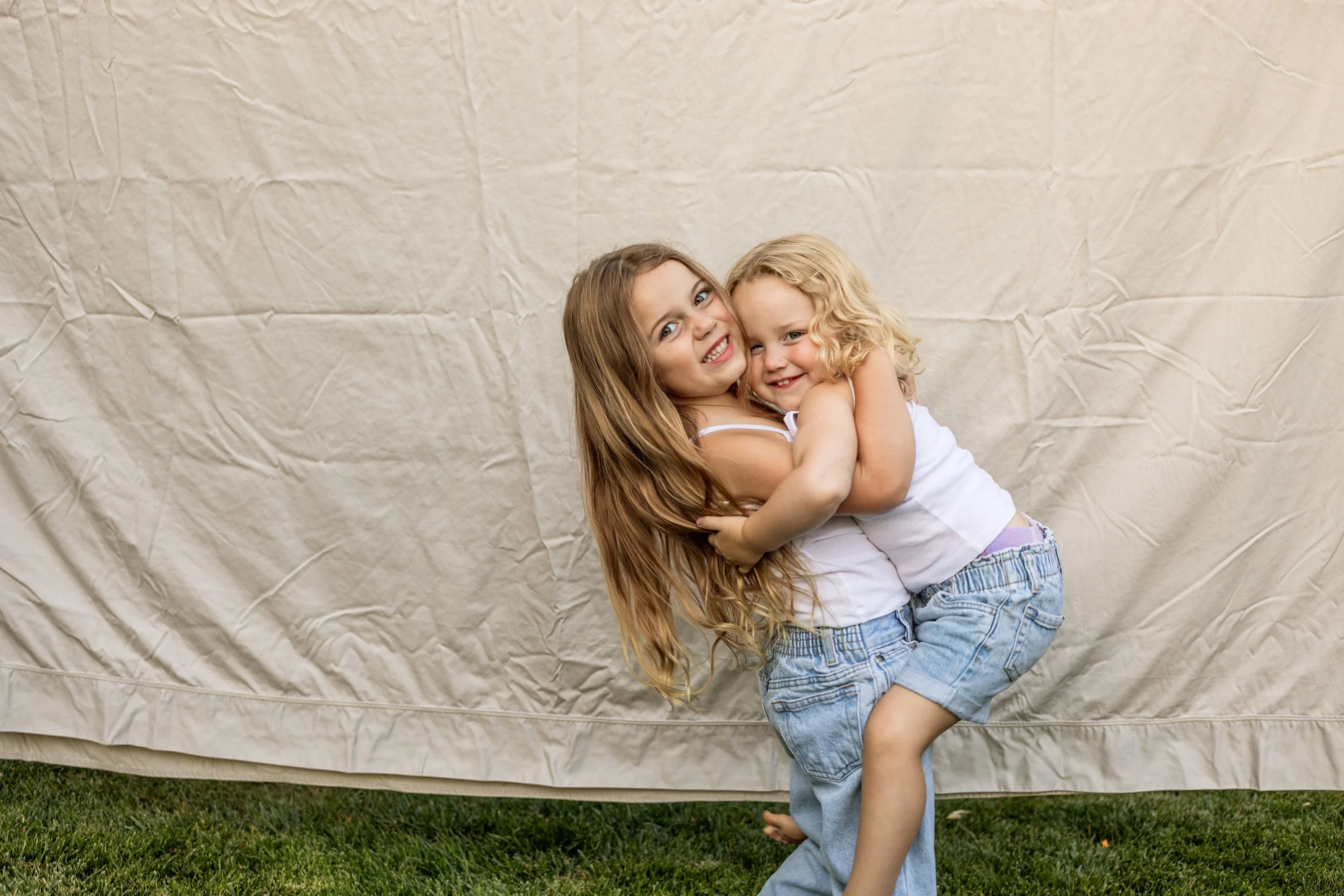 Two young girls with long hair hugging and smiling outdoors in front of a beige fabric backdrop, standing on grass.