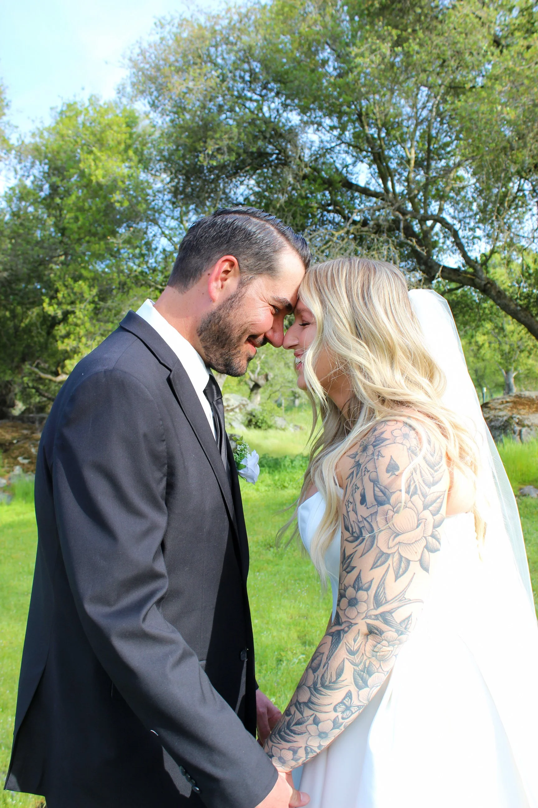 A bride and groom touching foreheads and smiling outdoors on their wedding day, holding hands, surrounded by trees and greenery.