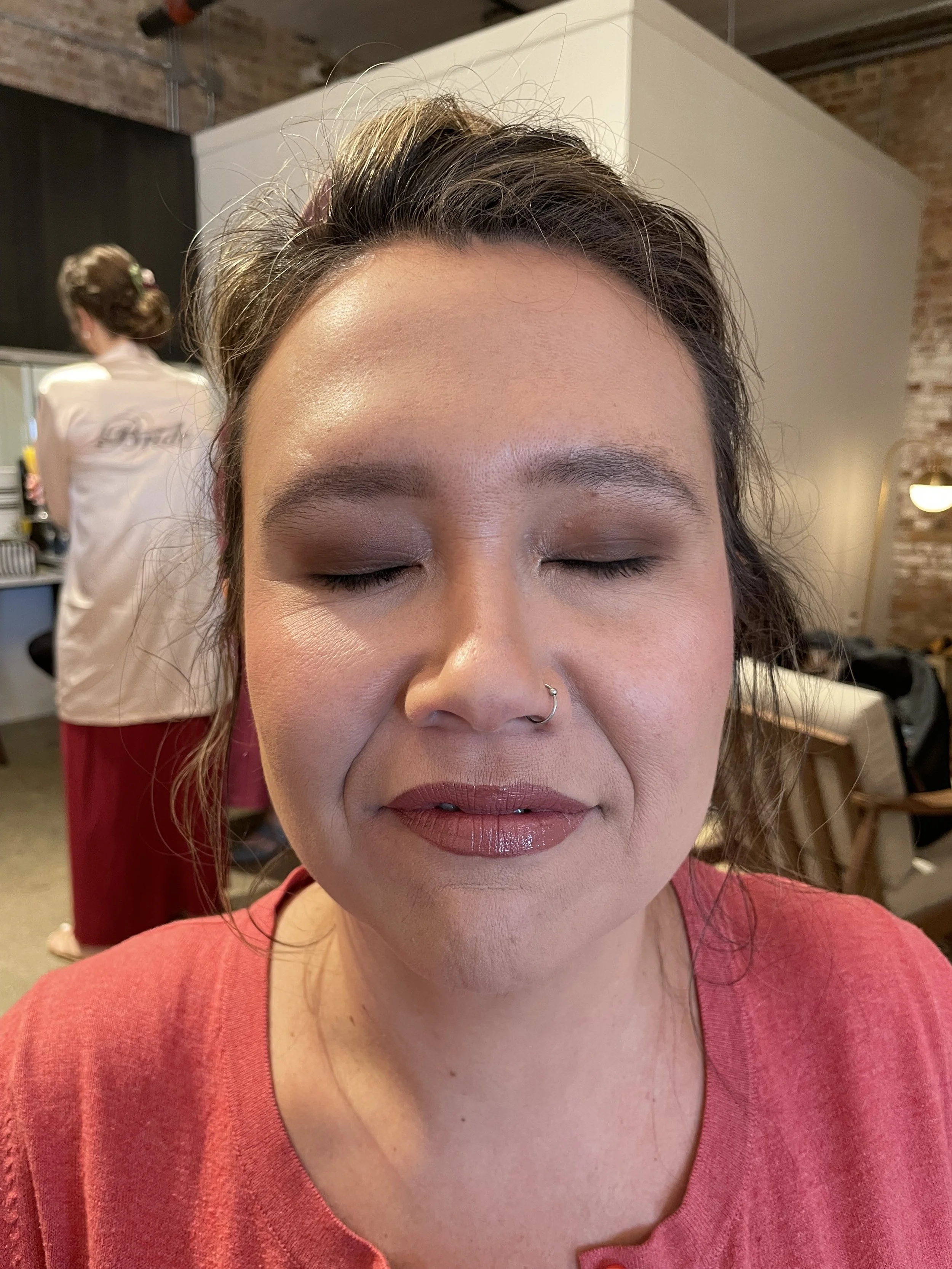 A woman with closed eyes and makeup, wearing a septum ring and a coral top, receiving a facial makeover in a salon with a stylist working in the background.