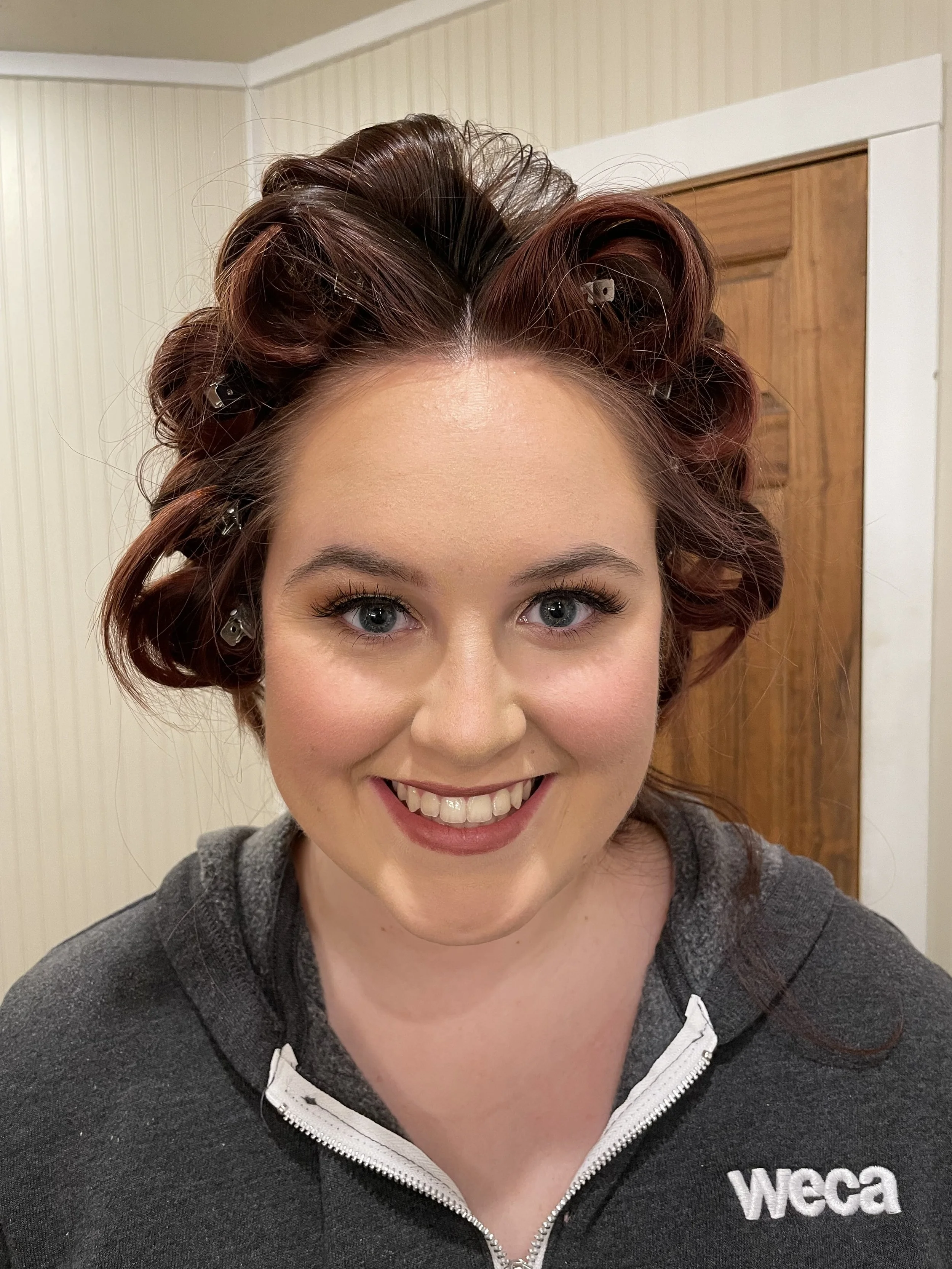 Bride with red hair in curlers, smiling, showing off her makeup.