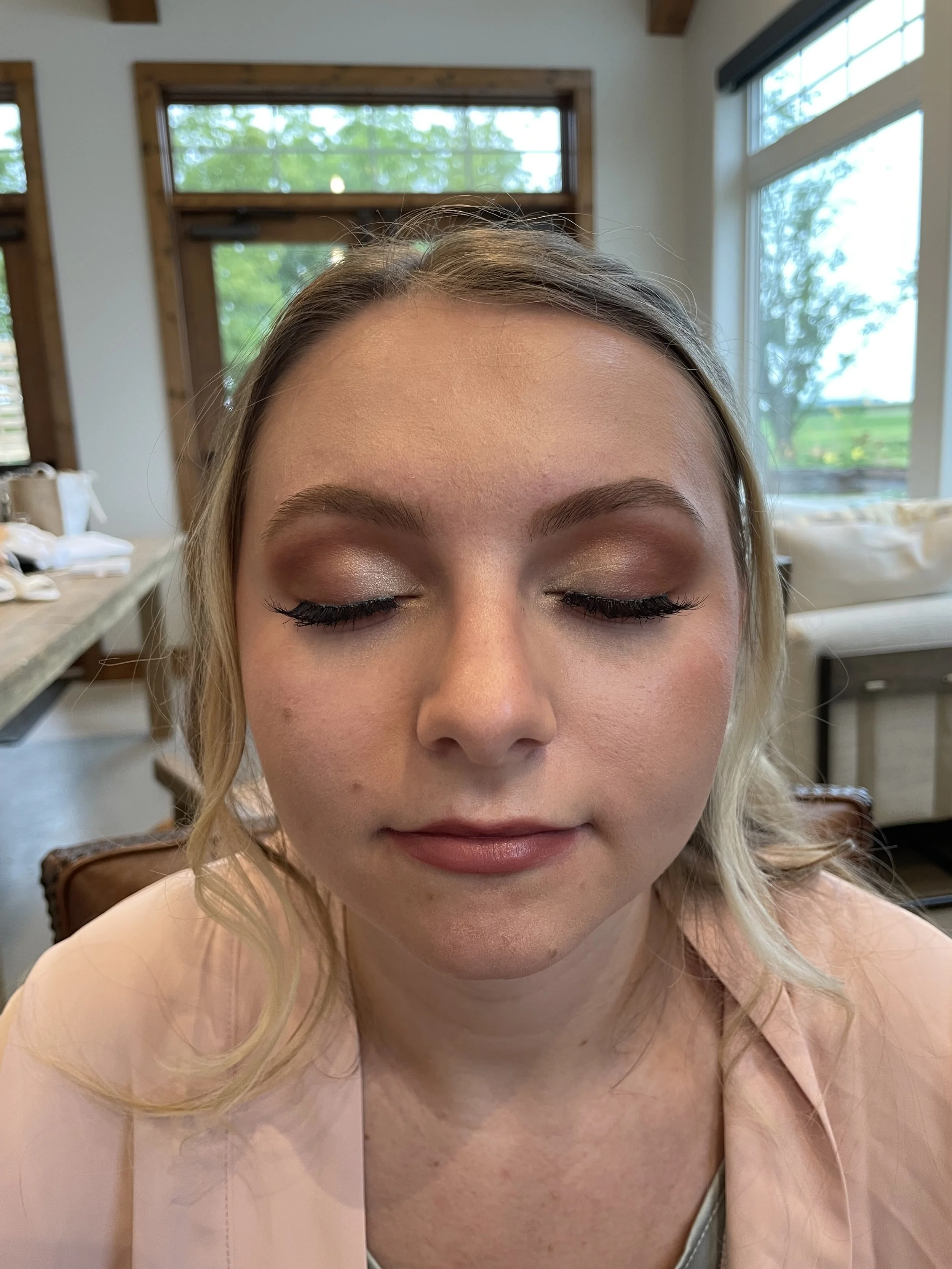 A young woman with blonde hair and makeup, eyes closed, sitting indoors in front of large windows showing green trees outside.