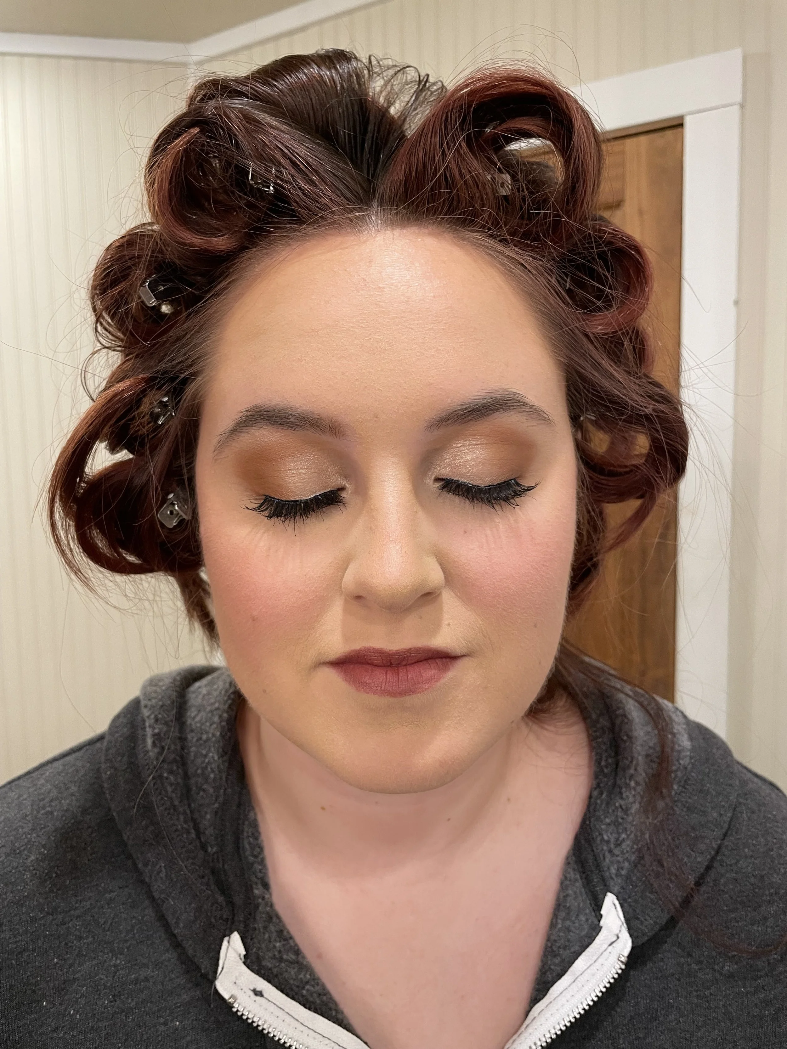 Bride with closed eyes, wearing makeup, and curlers in her hair, dressed in a gray hoodie.