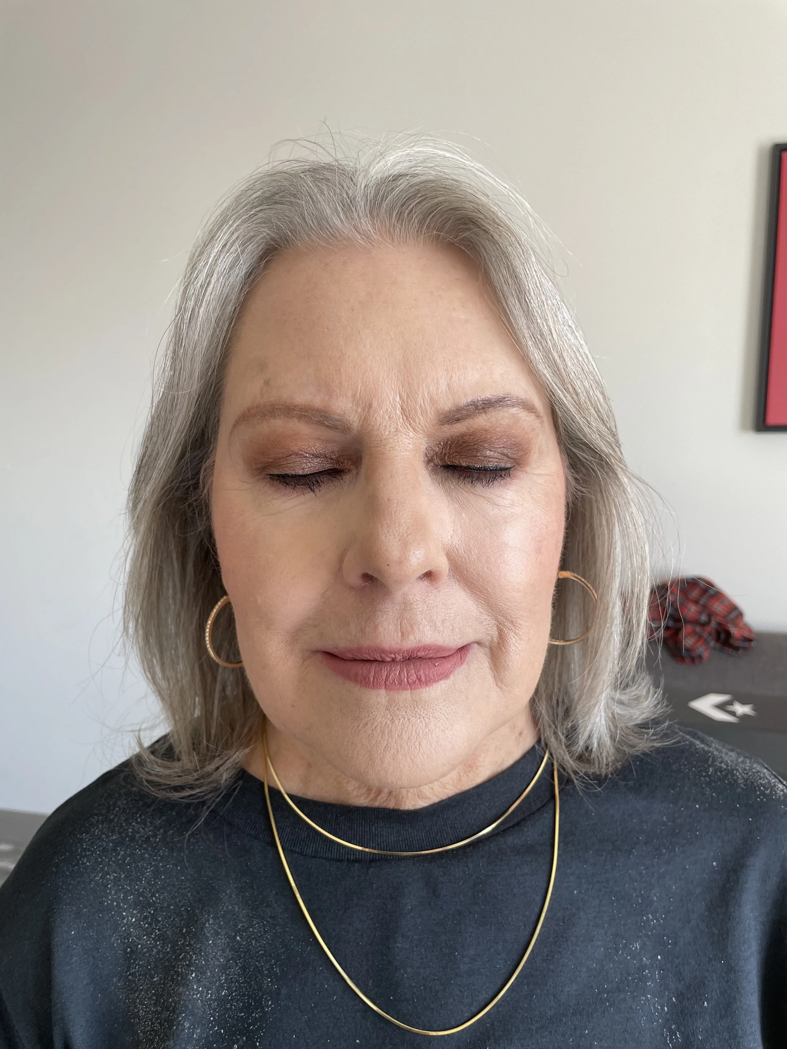 A close-up of a woman with gray hair, closed eyes, wearing gold hoop earrings, two gold necklaces, and a black shirt with glitter details, standing against a plain background.