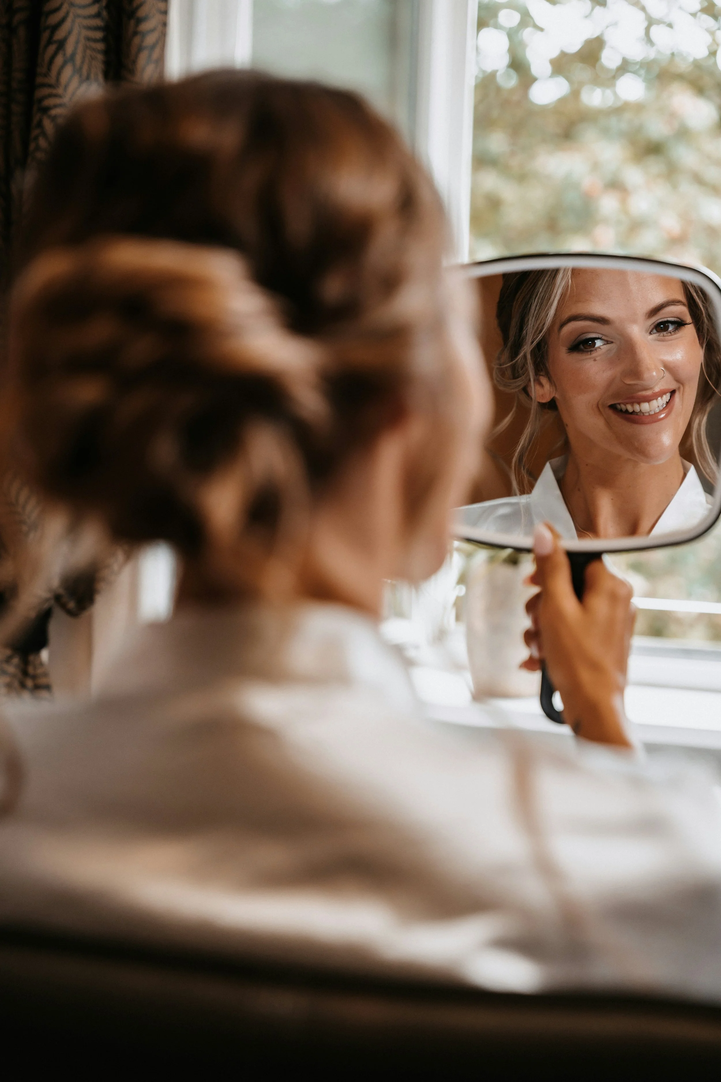 A woman with curled hair smiling as she looks into a small handheld mirror, with her reflection showing her face.