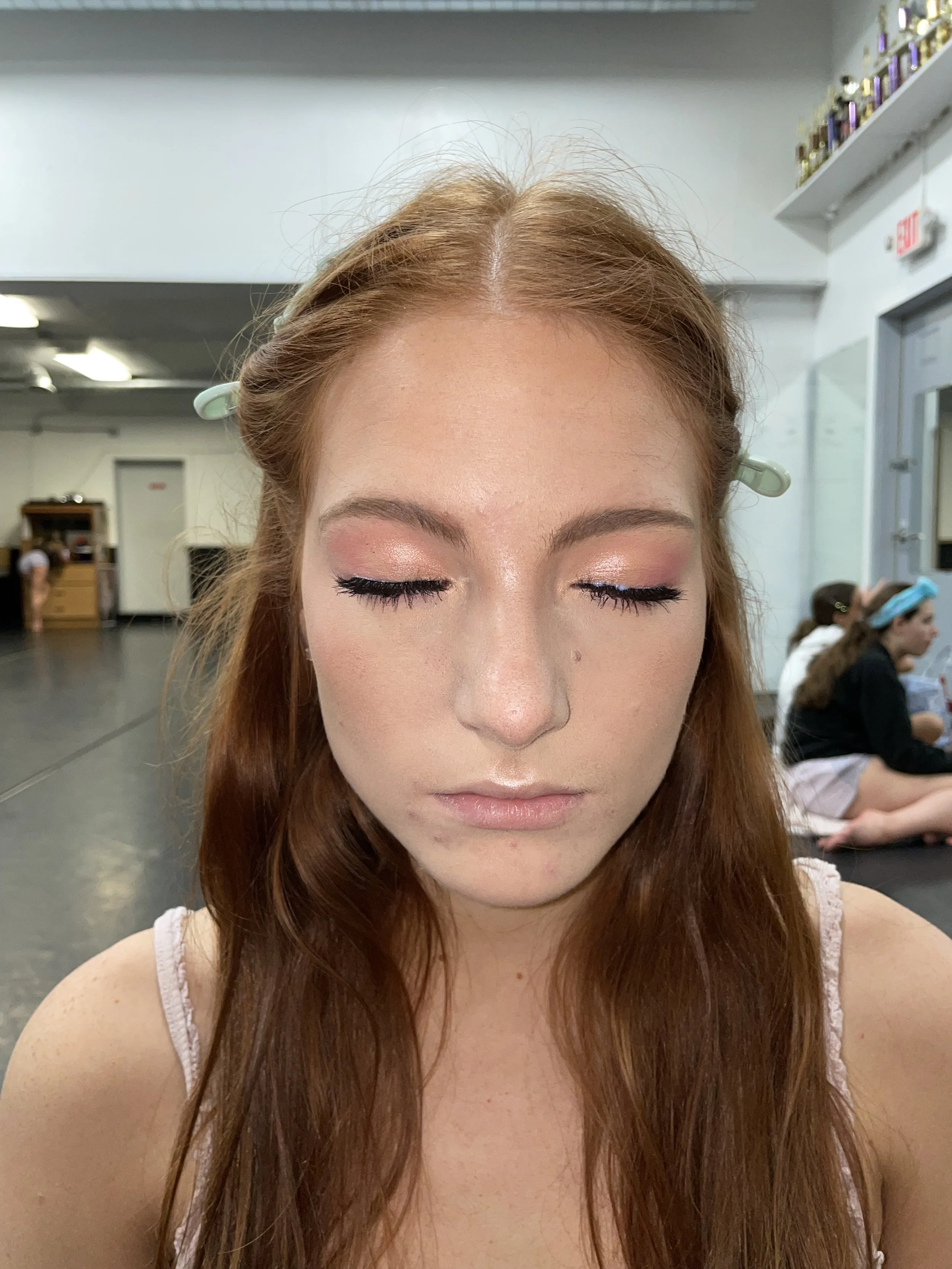 A young woman with closed eyes, red hair, and pink-toned makeup getting ready for prom in a dance studio.