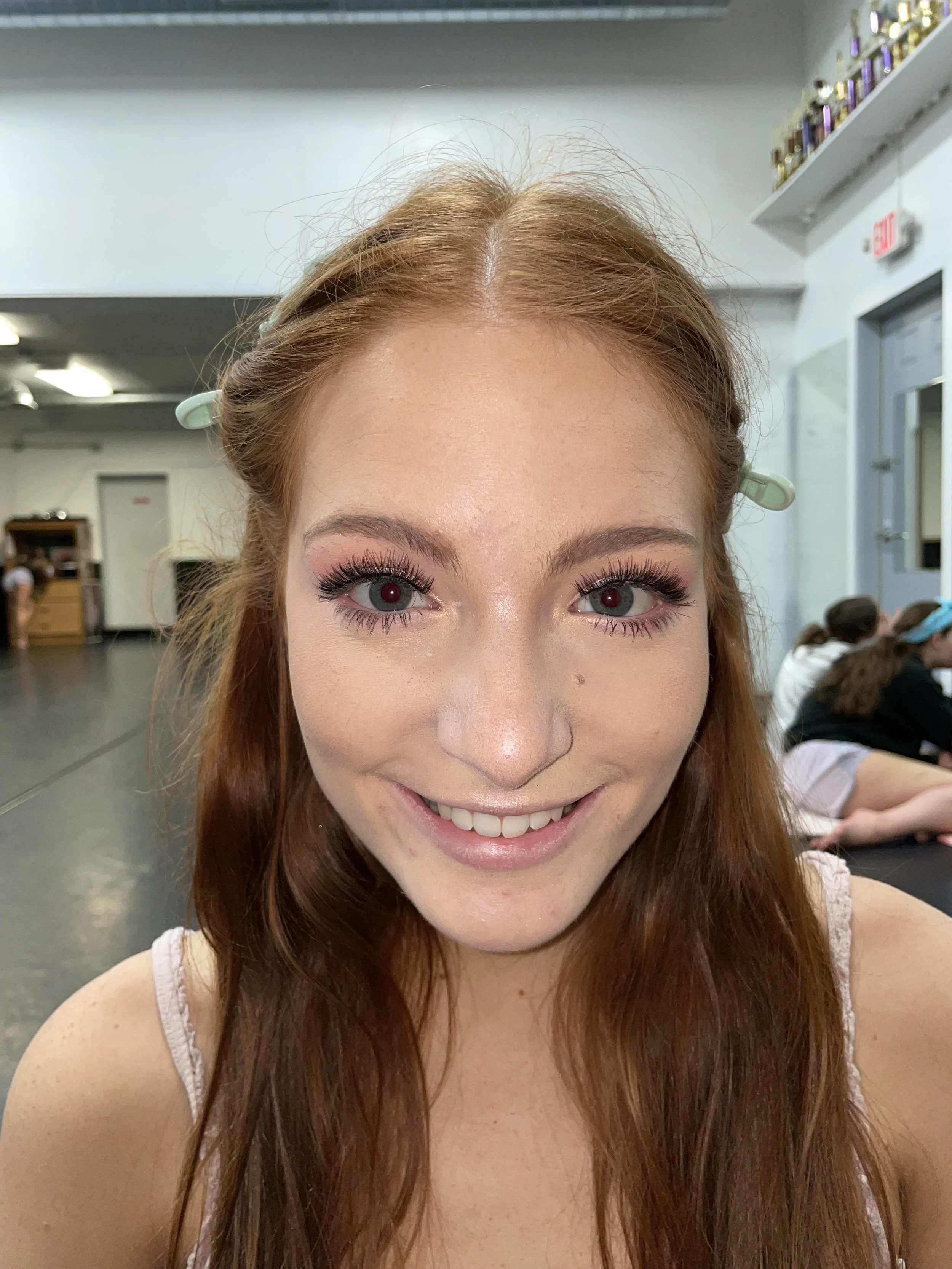 A young woman smiling at the camera with red hair, blue eyes, and pink-toned prom makeup.