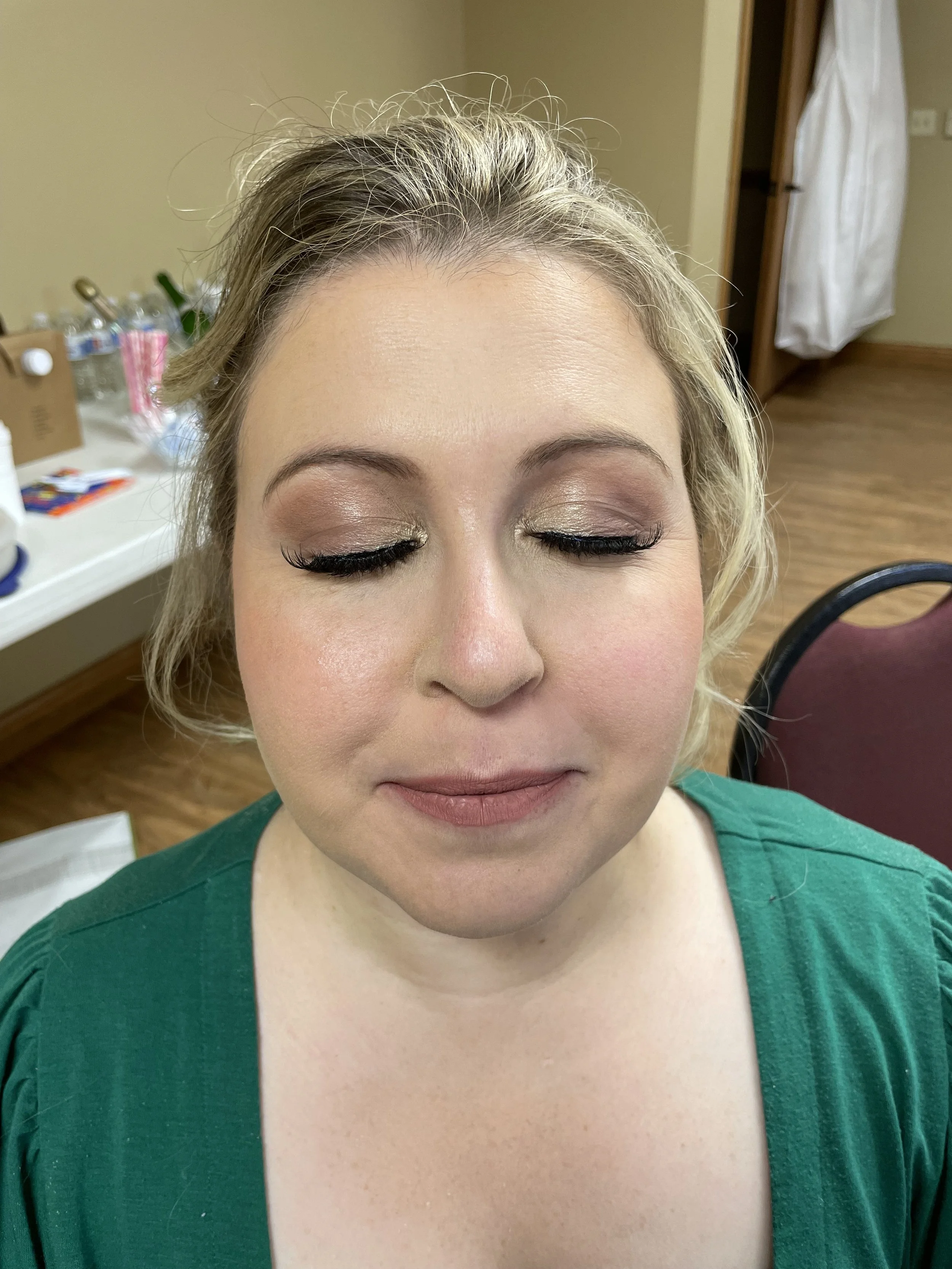 Close-up of a woman with blonde curly hair and closed eyes, showing eye makeup and lipstick, in a bridal getting-ready room.
