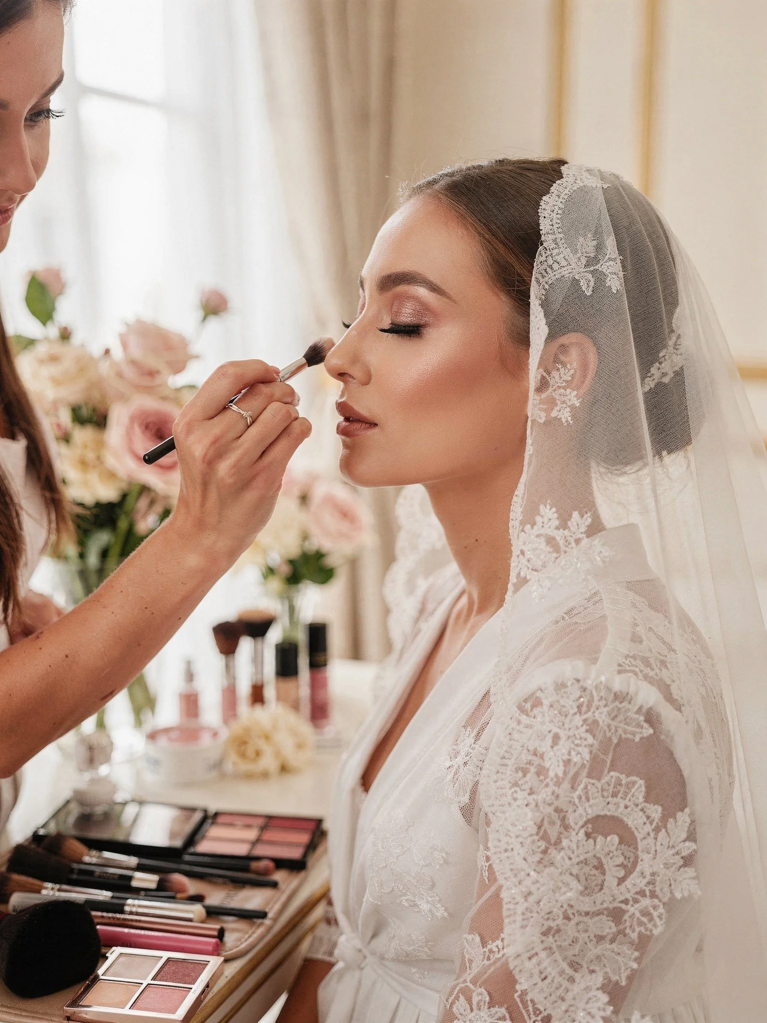 Bride with lace veil having makeup applied by a makeup artist in a floral-filled room.