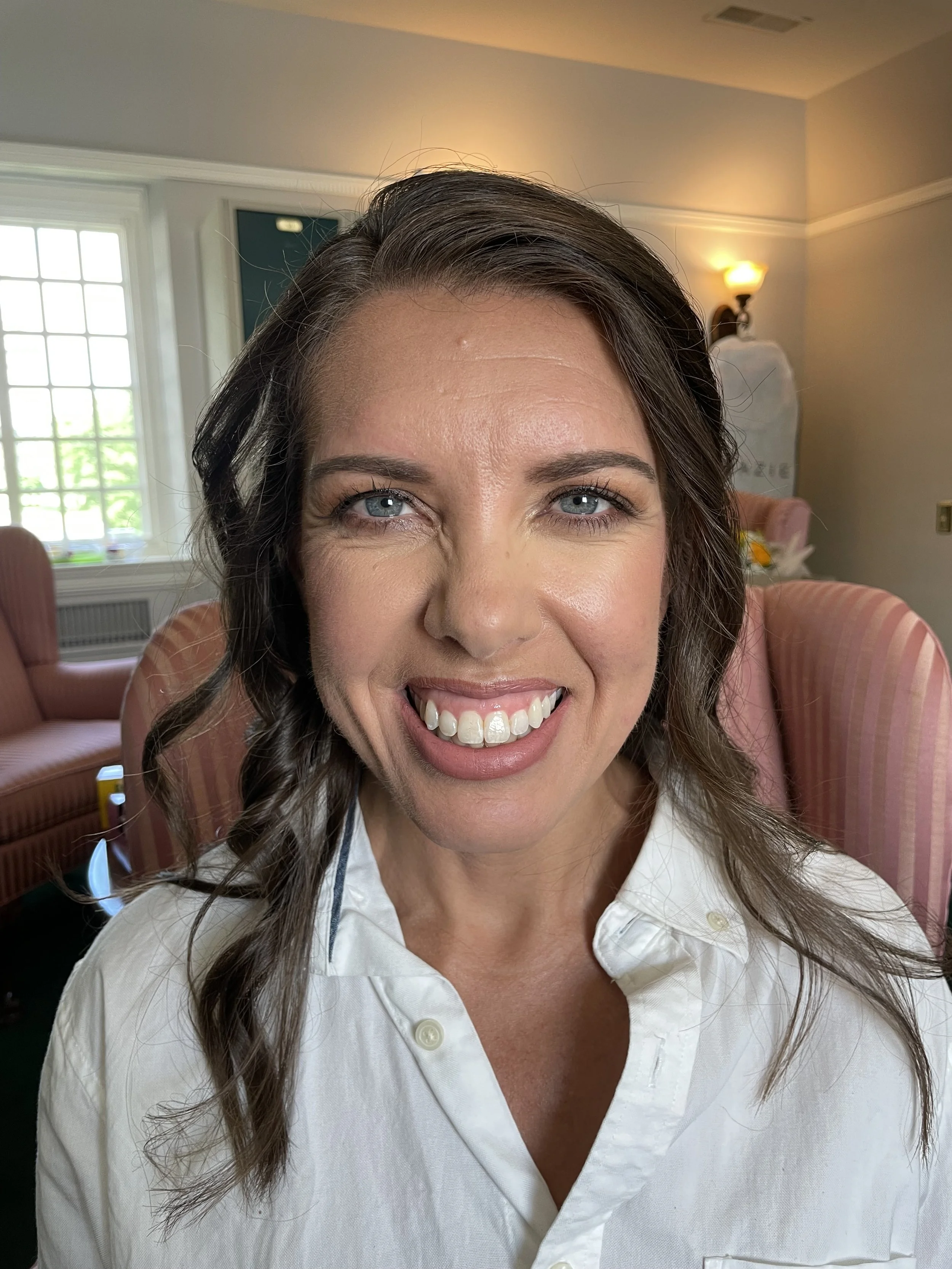 Close-up of a smiling woman with brown hair and blue eyes, wearing a white shirt, in a well-lit indoor room with pink chairs and a window in the background.