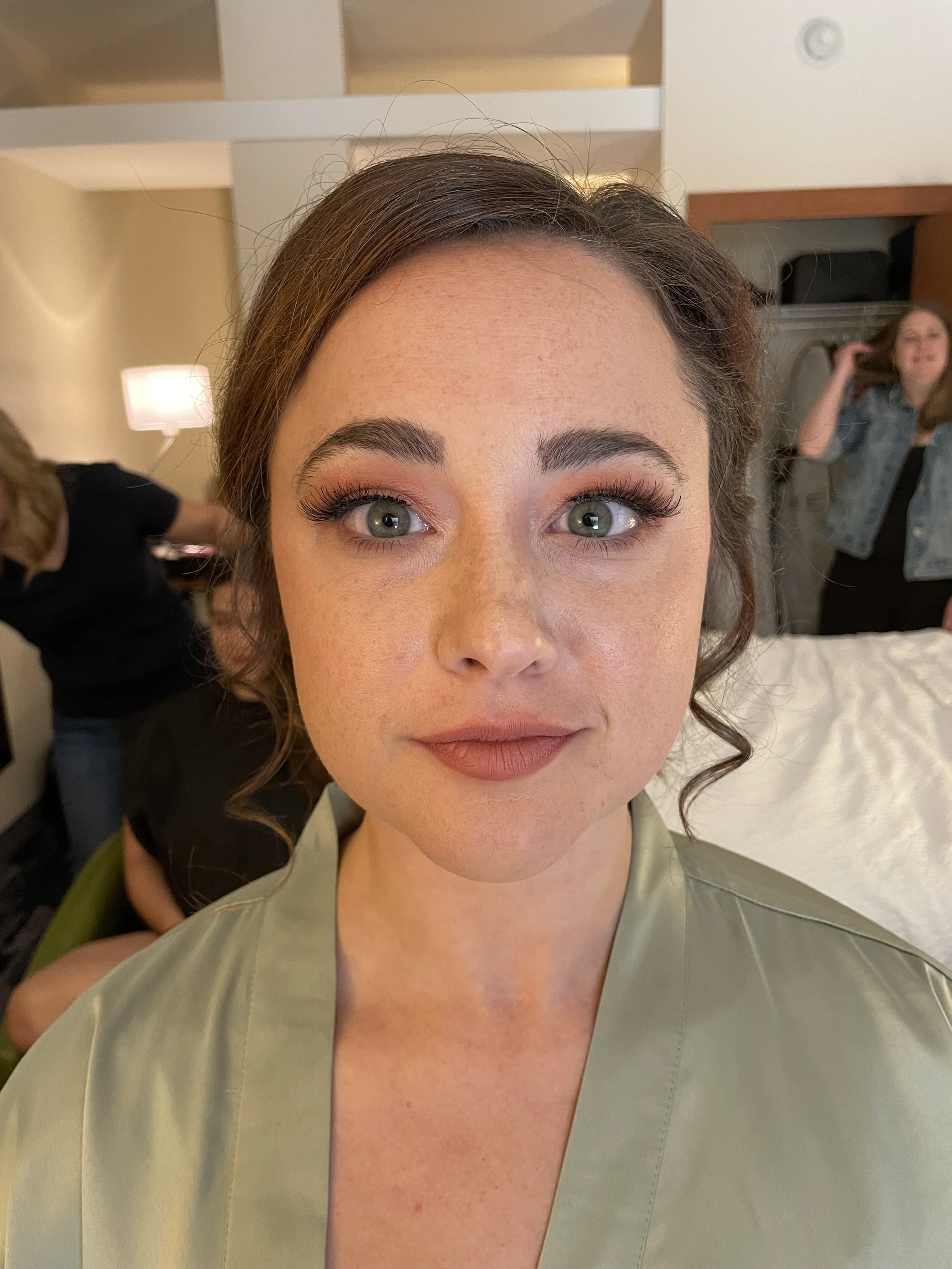 Close-up of a woman with makeup in a hotel room, with two people in the background, one sitting and the other adjusting her hair.