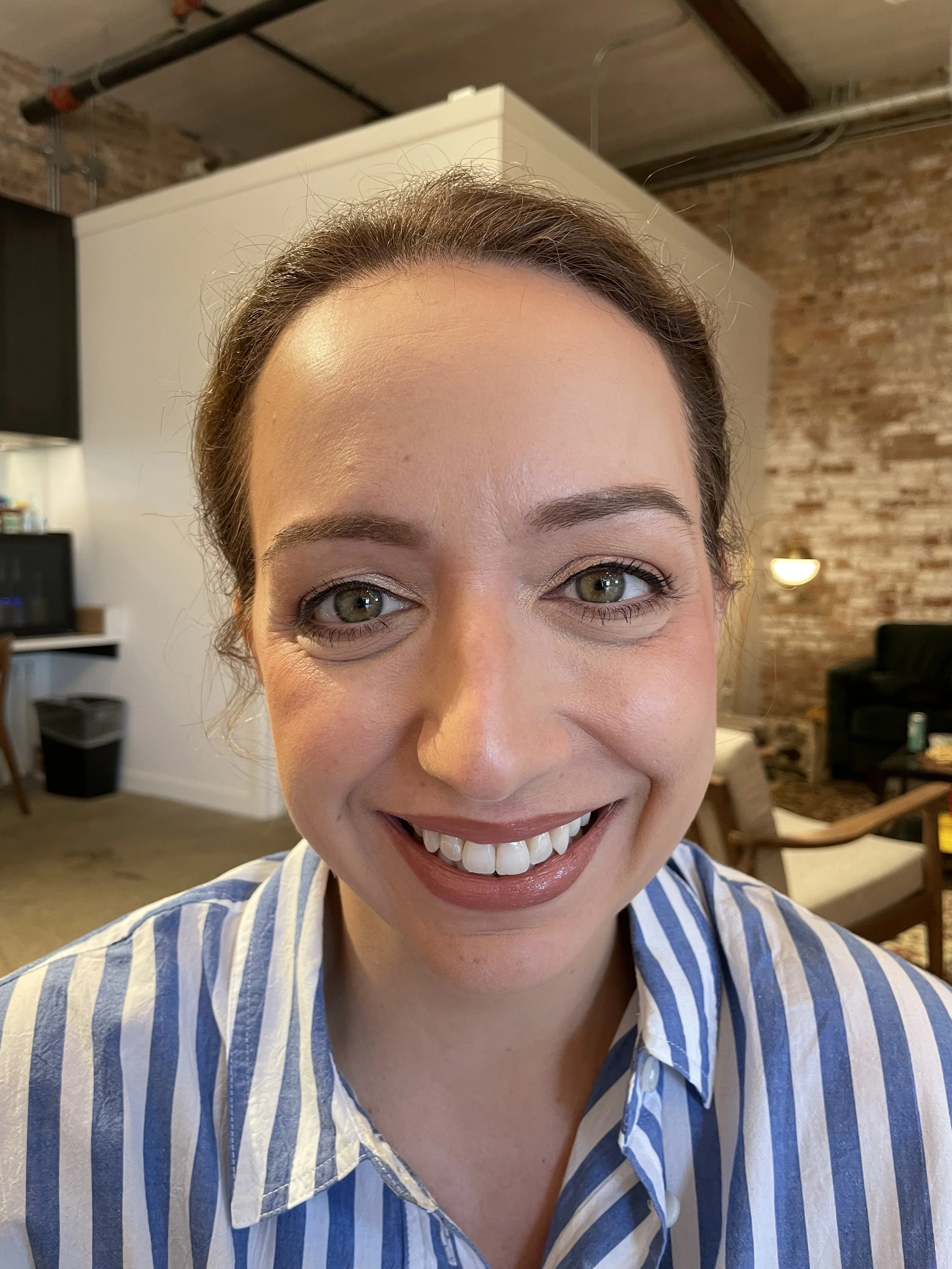 Close-up of a smiling woman with natural makeup, light brown hair and green eyes, wearing a blue and white striped shirt.