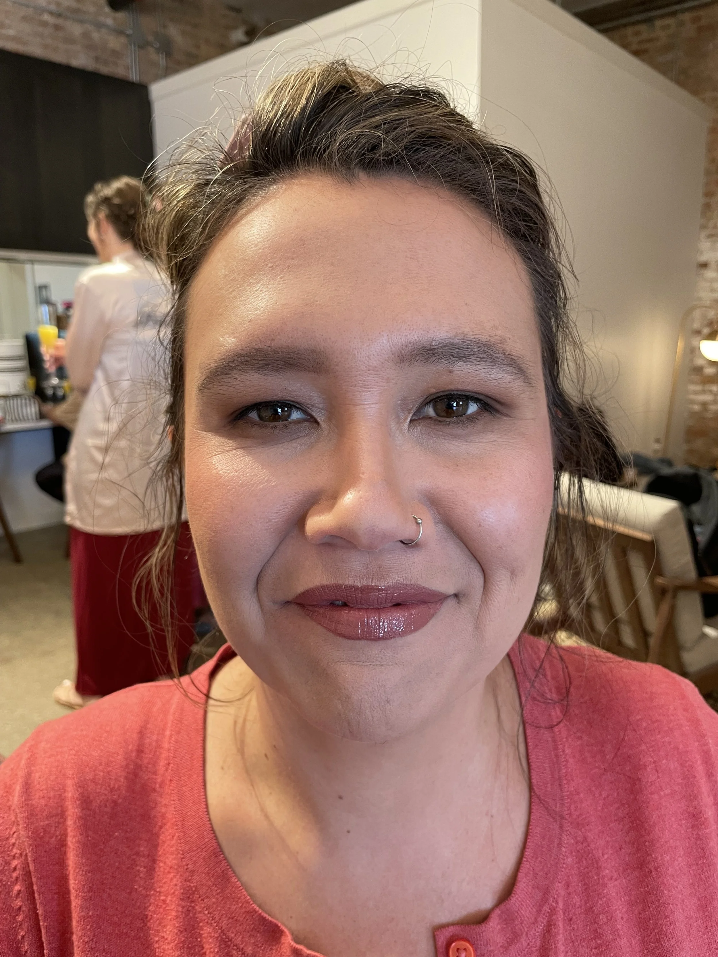 Close-up of a smiling woman with dark hair, brown eyes, a nose ring, wearing a pinkish-red top, in a cozy indoor setting.
