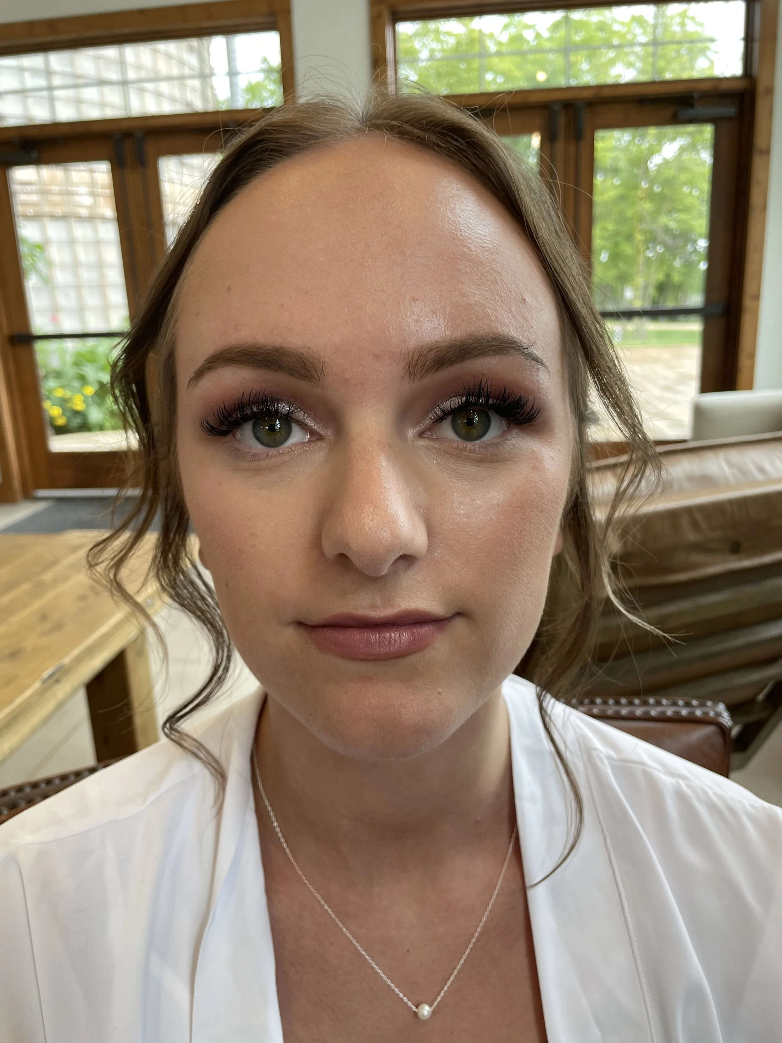 A woman with wavy brown hair and light skin looking at the camera, wearing makeup with false eyelashes, a white top, and a delicate necklace with a pearl pendant, seated in a bright room with large windows showing green outdoor scenery.