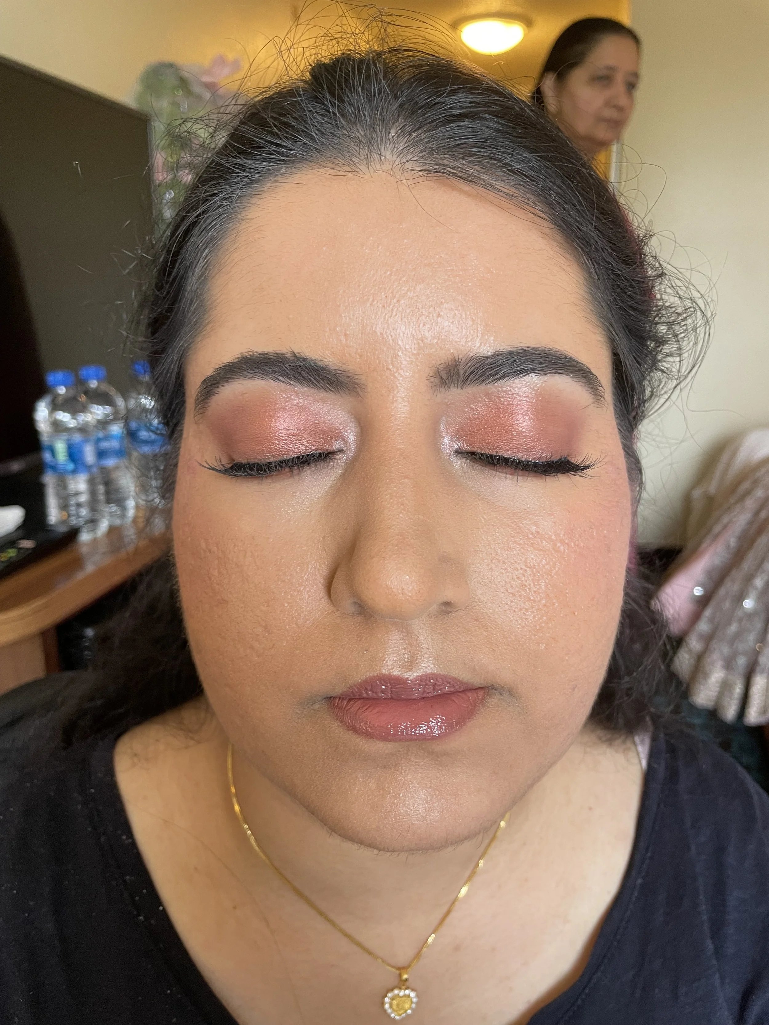Close-up of a woman's face with closed eyes, showcasing makeup, in a room with a woman in the background and bottles of water on a table.