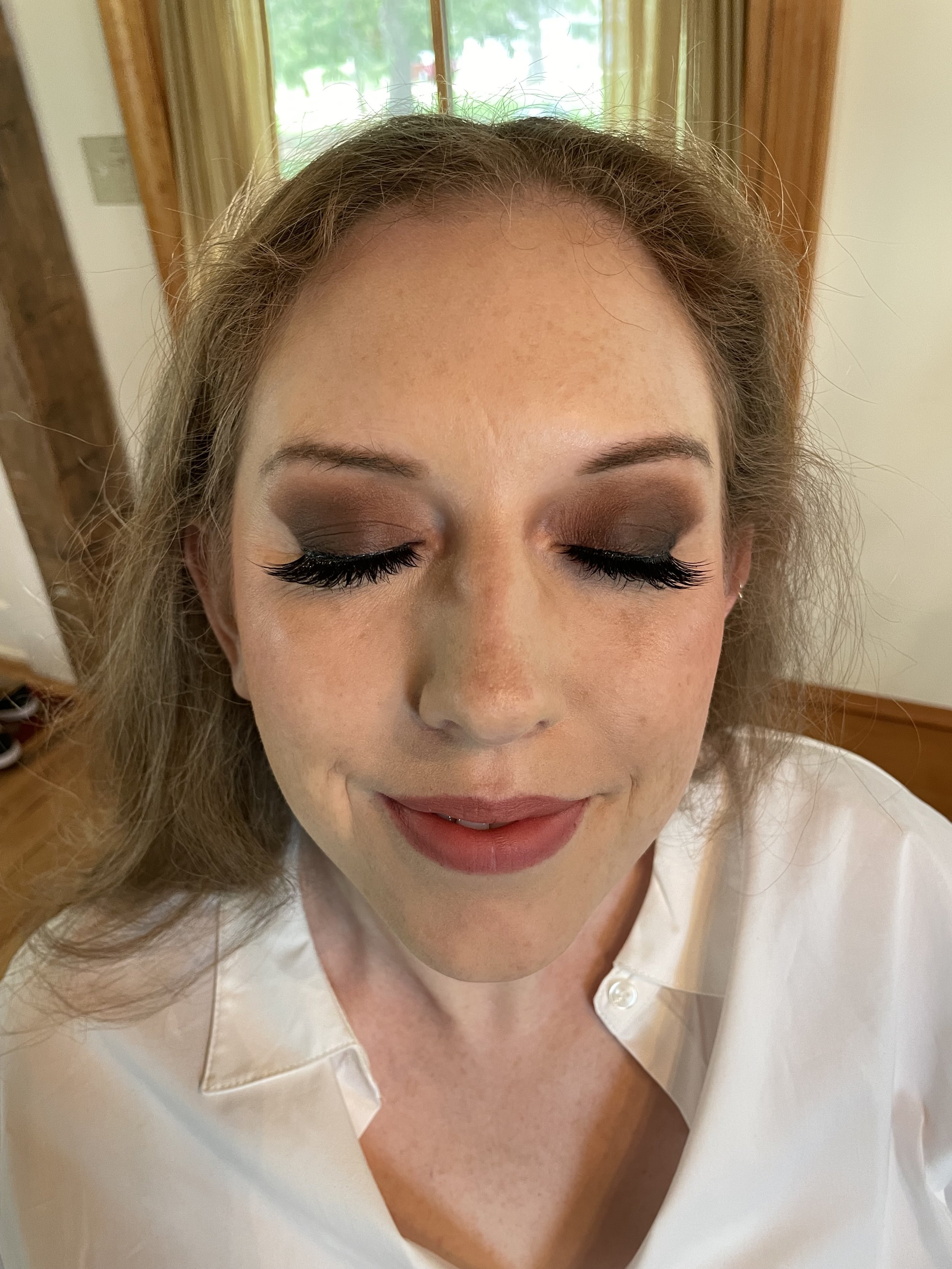 Close-up of a woman with closed eyes showing smoky eye makeup and long eyelashes, smiling slightly. She has light skin with freckles, and her hair is wavy.