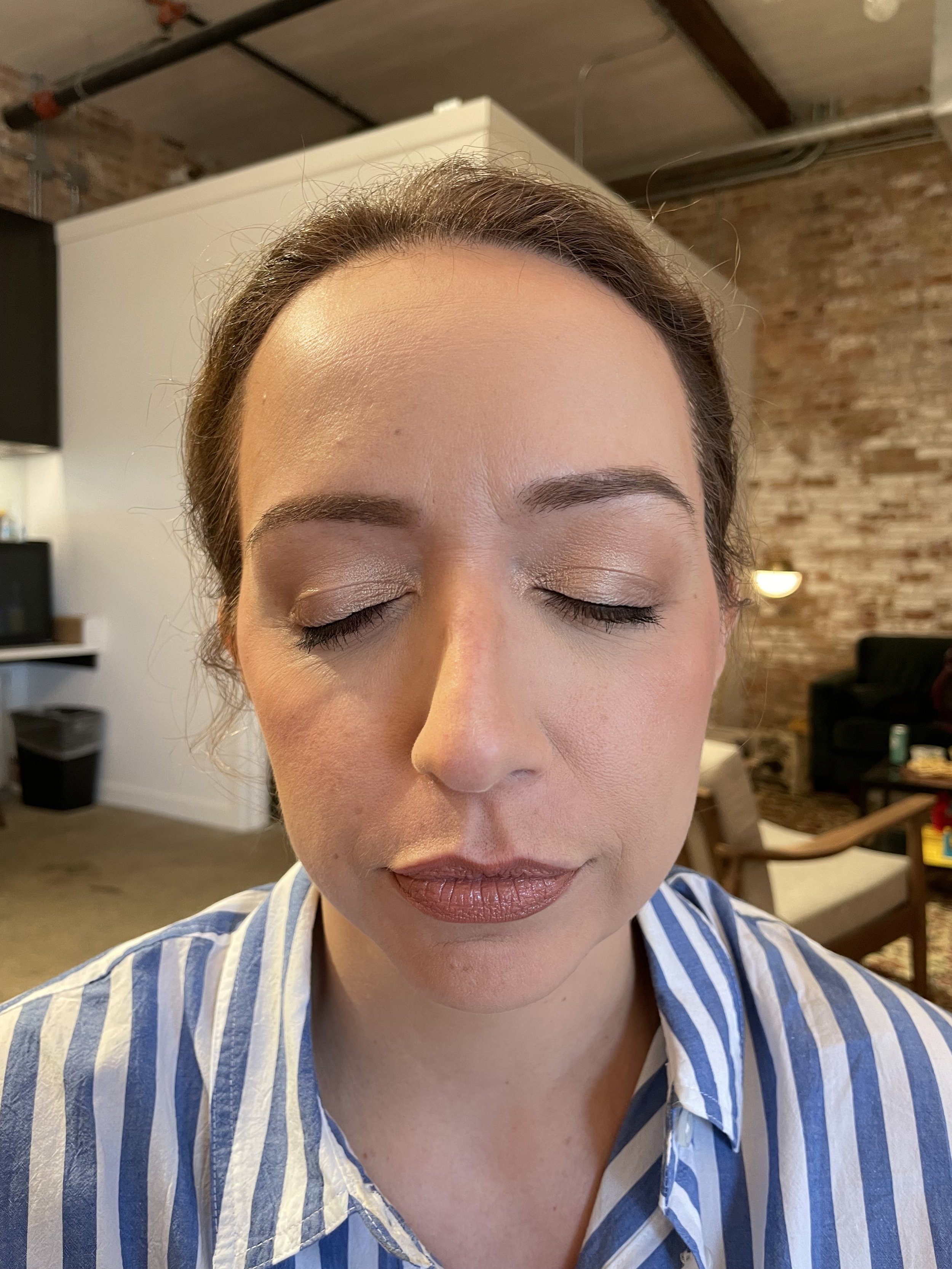 Close-up of a woman with closed eyes and natural makeup, wearing a blue and white striped shirt, in an indoor setting.