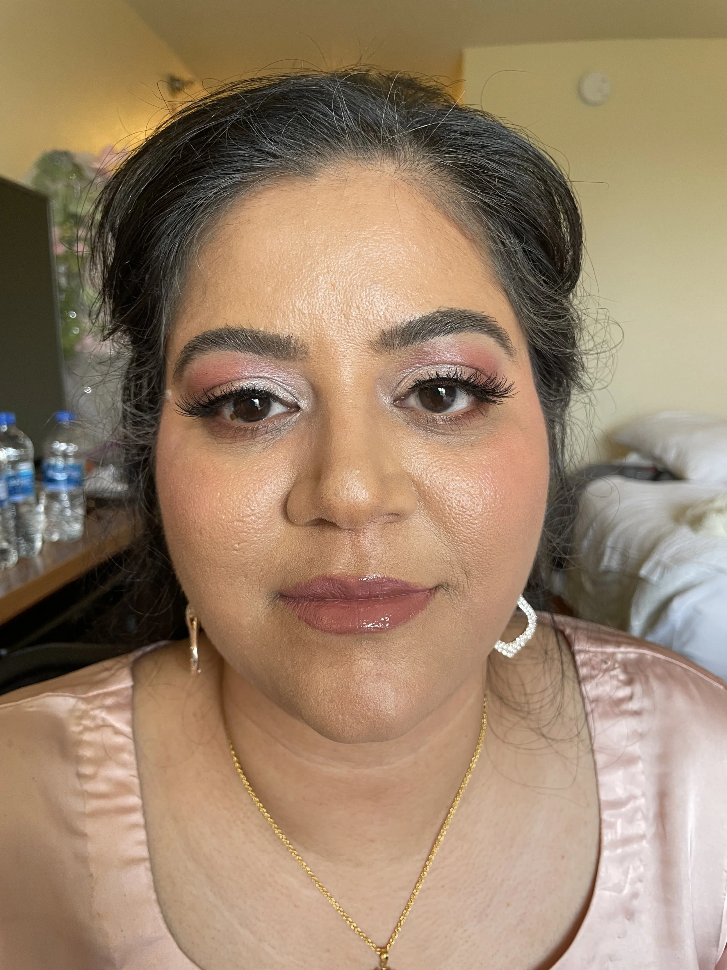 A woman with dark hair styled back, wearing makeup with pink and silver eyeshadow, glossy pink lipstick, and hoop earrings. She has a gold necklace and is dressed in a light pink satin top.