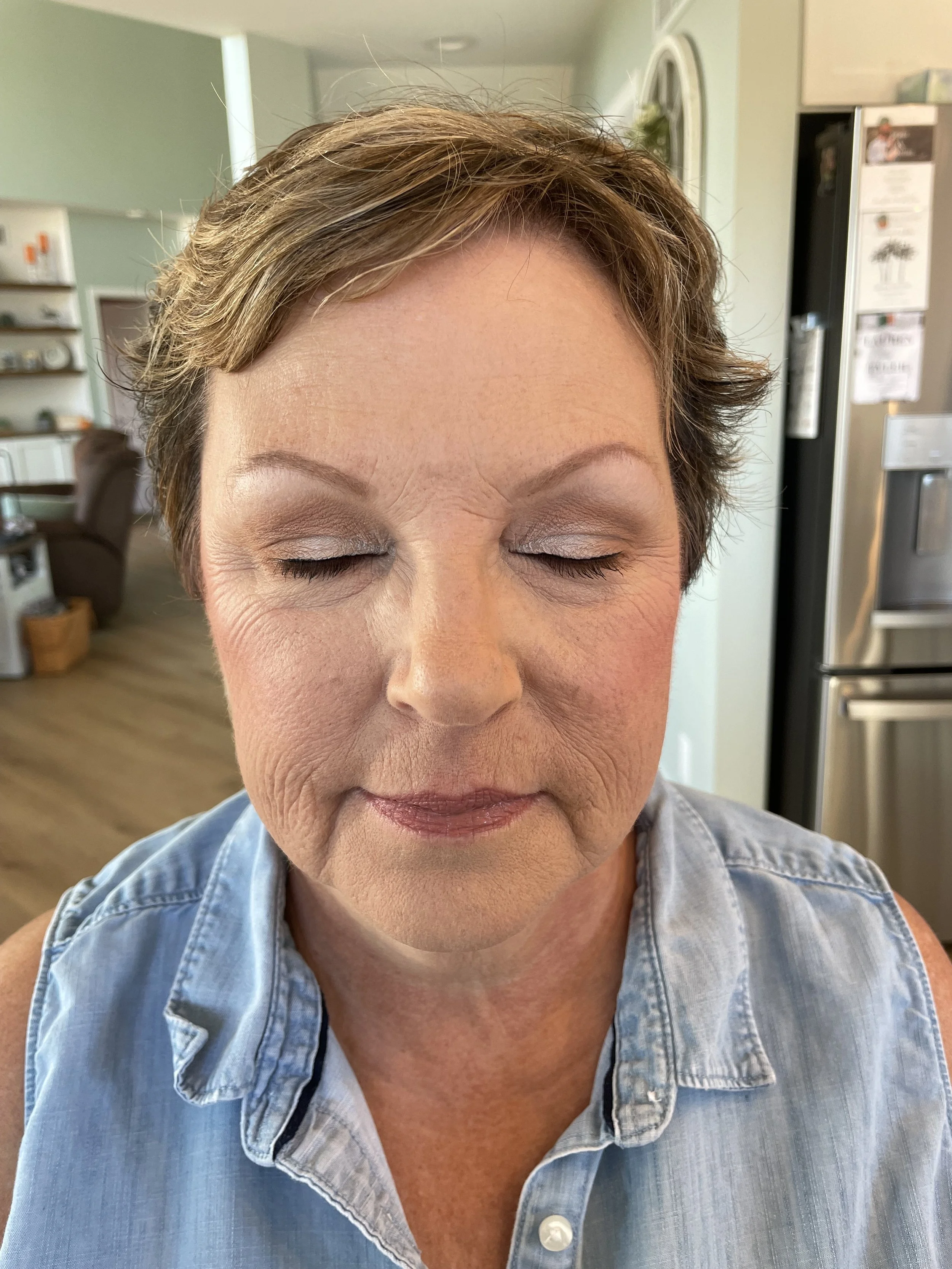An older woman with short brown hair and closed eyes, wearing a sleeveless denim shirt, in a home setting with kitchen appliances and living room furniture in the background.