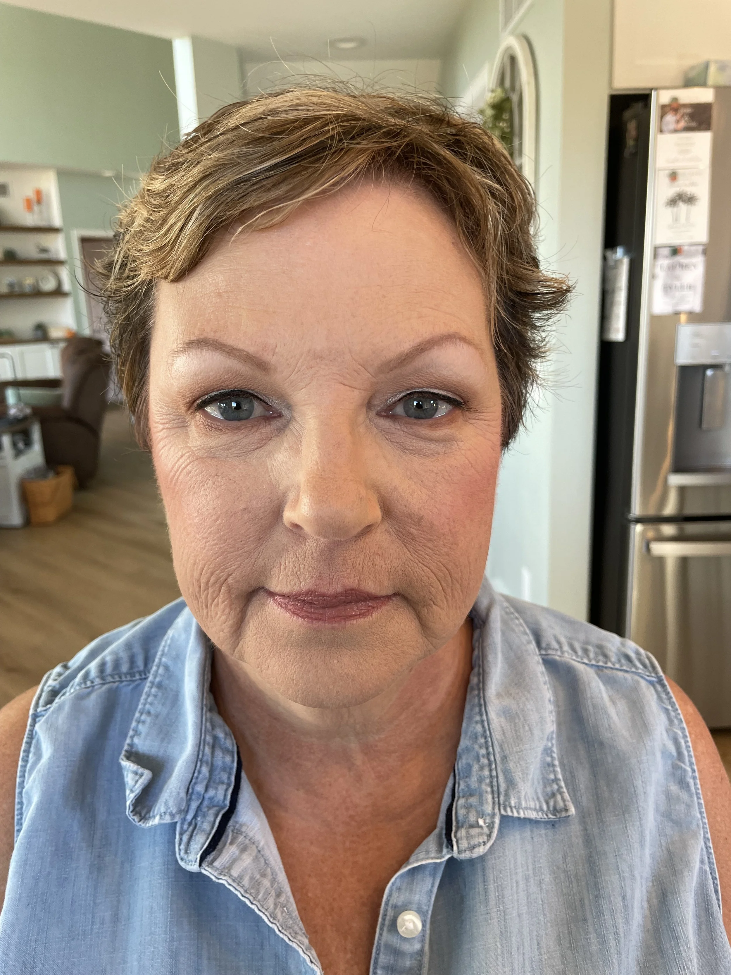 Close-up of a middle-aged woman with short hair in a kitchen, wearing a sleeveless denim shirt.