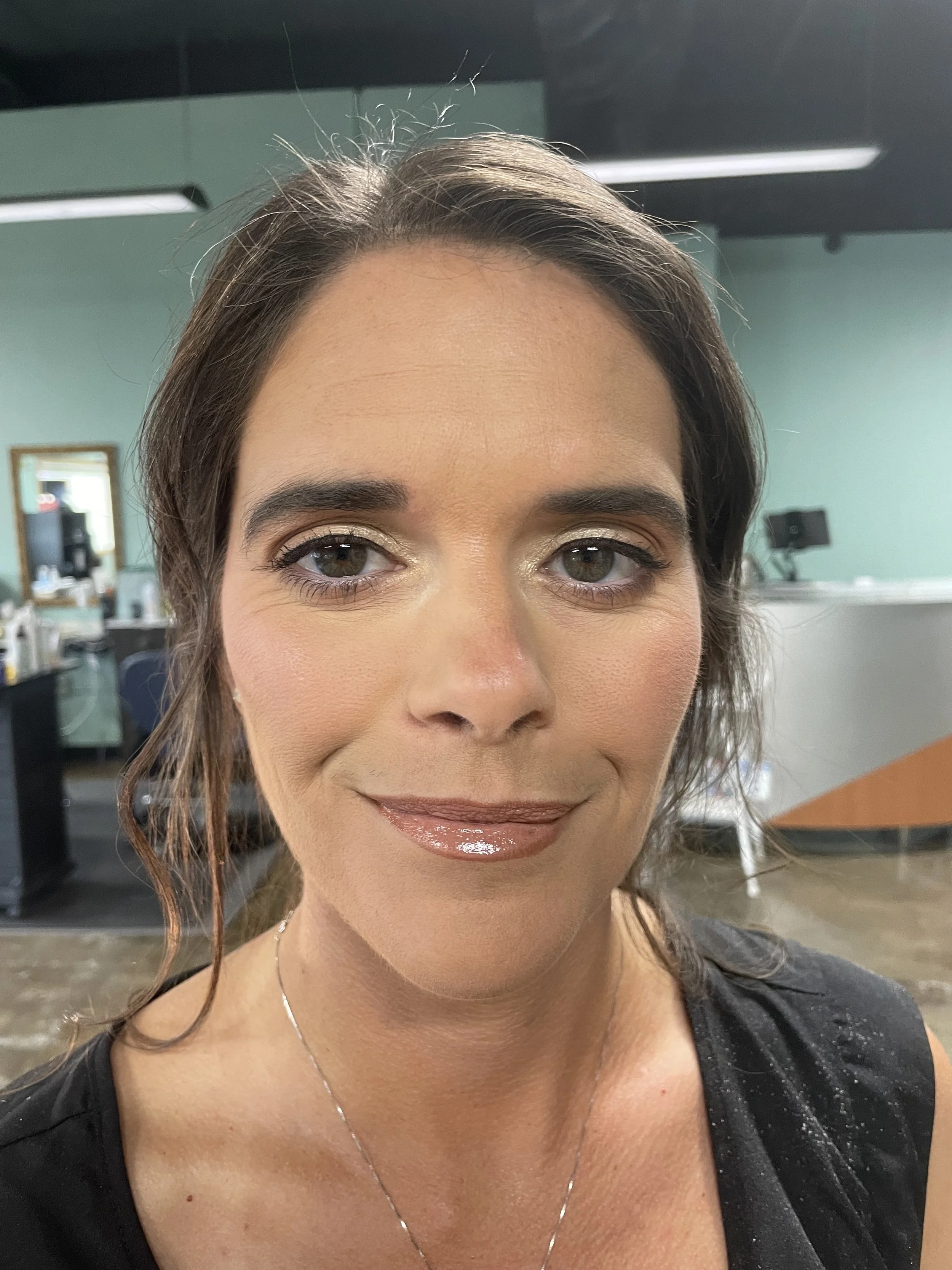 Close-up of a woman with brown hair, wearing makeup, a black top, and a silver necklace, smiling in a salon or studio background.
