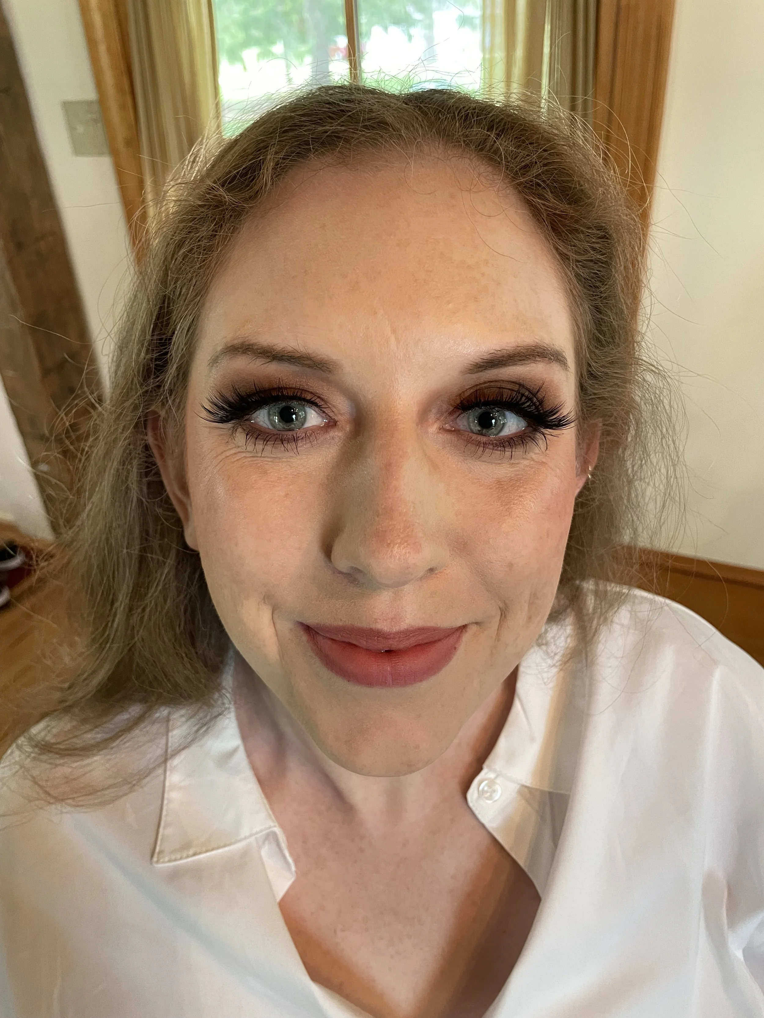 A close-up of a bride with light skin, blue eyes,  smoky eye makeup, and long eyelashes, smiling at the camera. She has light skin and brown wavy hair.