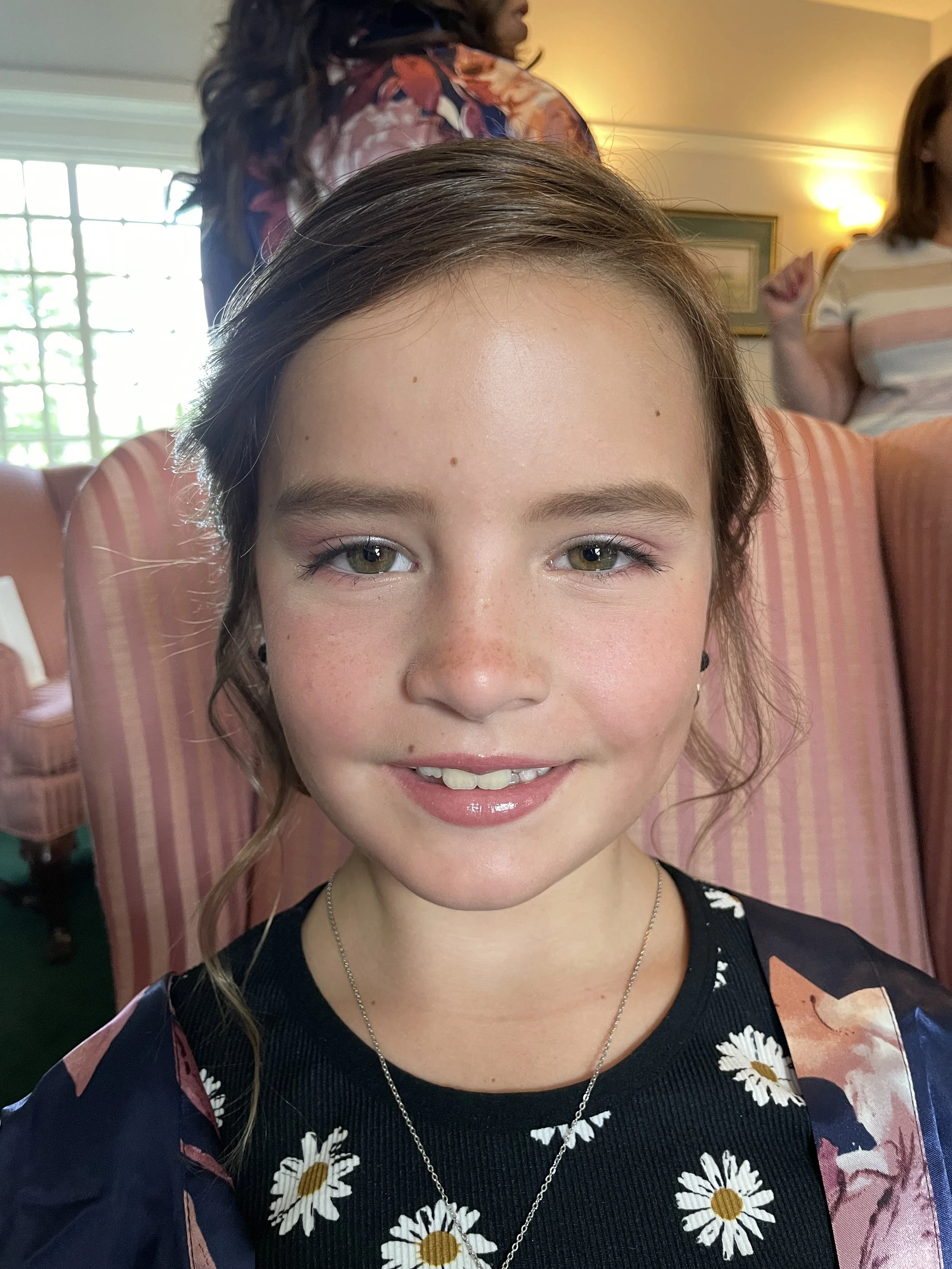 A young flower girl with light skin, light brown hair, and green eyes smiling at the camera, wearing a black shirt with daisy flowers and a silver necklace, seated indoors.