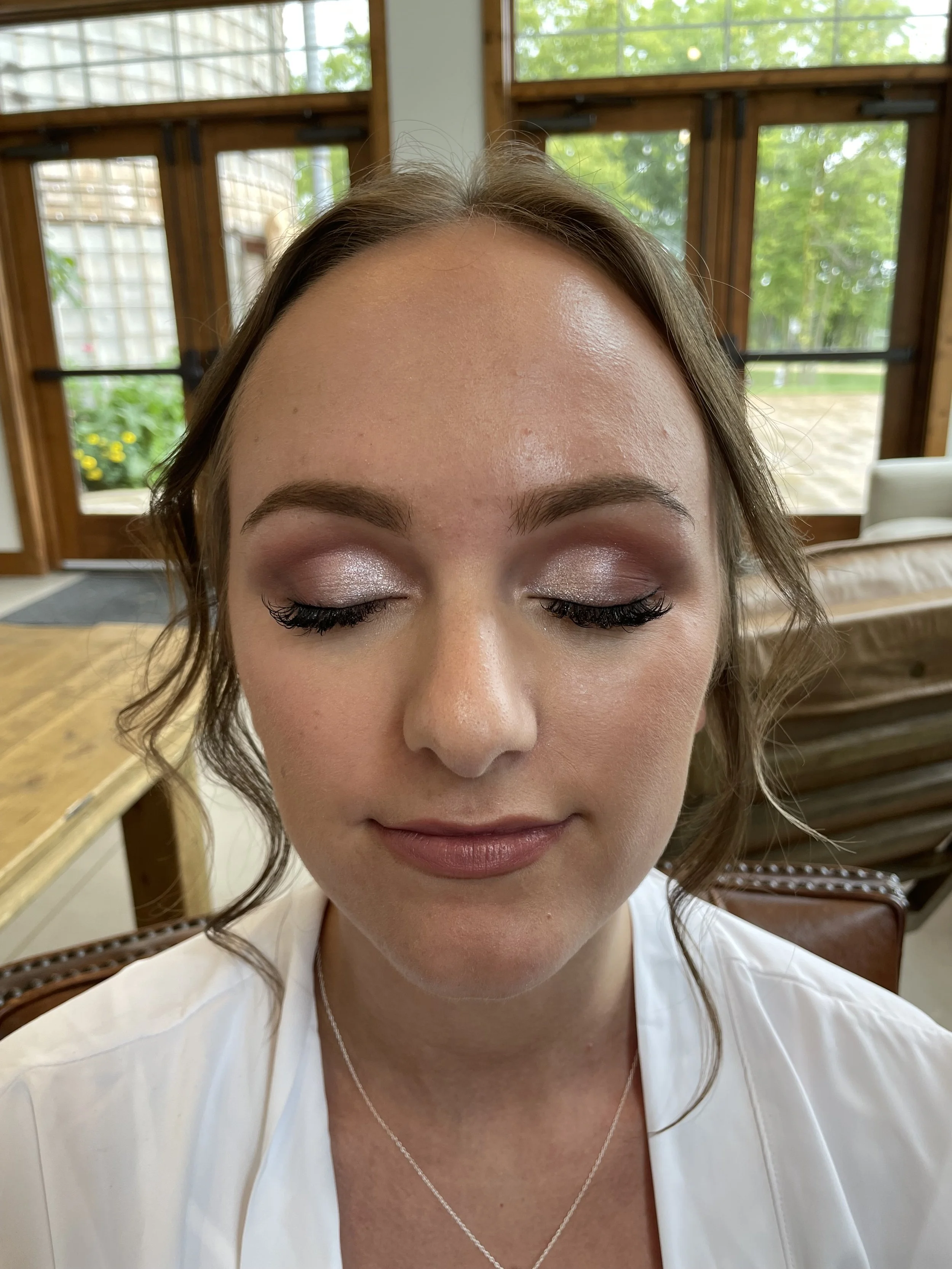 A woman with eyes closed, showing her makeup, in a room with large windows and green trees outside.
