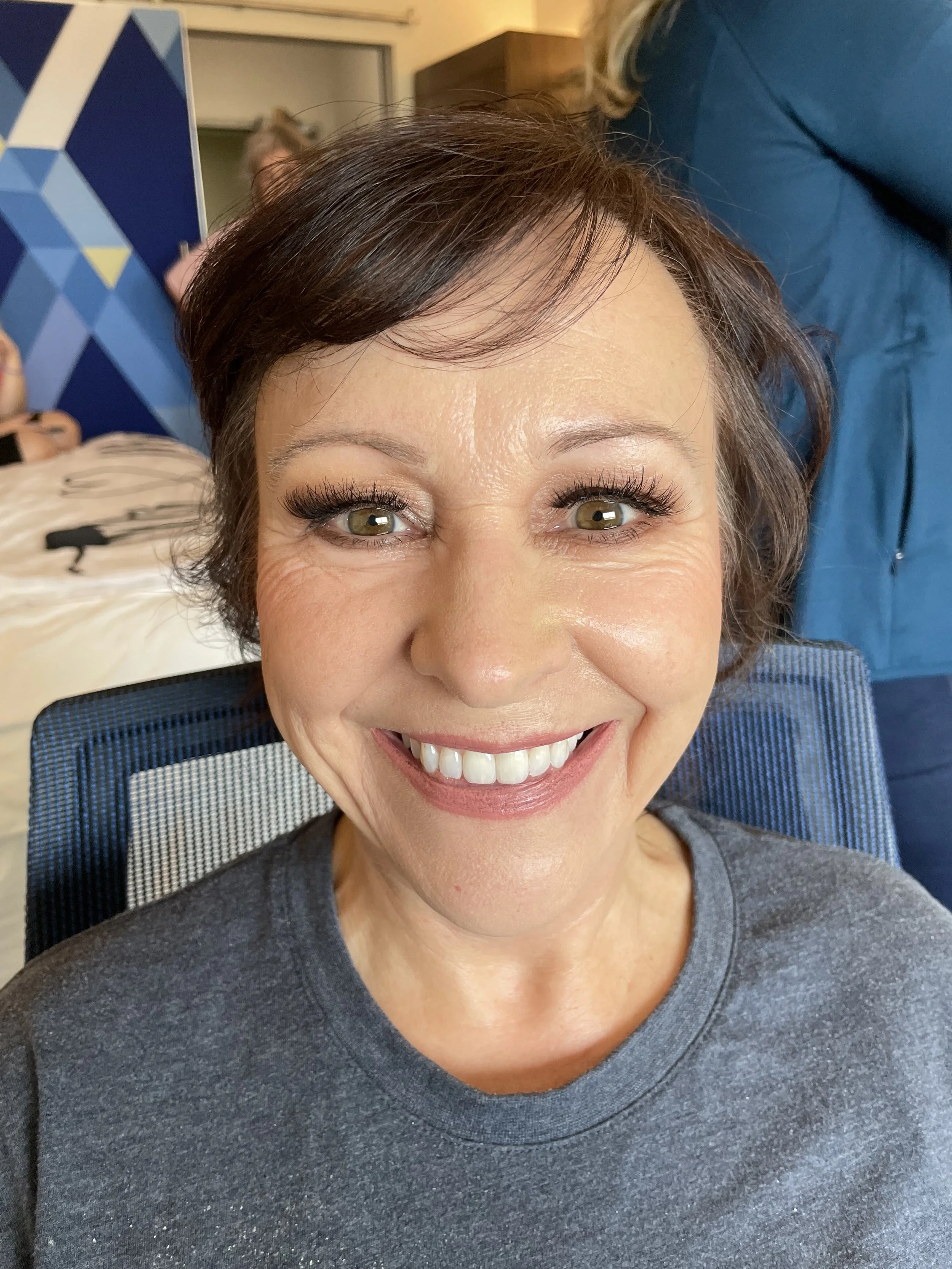 Close-up of a mother of the bride/groom with short brown hair and green eyes, smiling at the camera showing off her makeup.