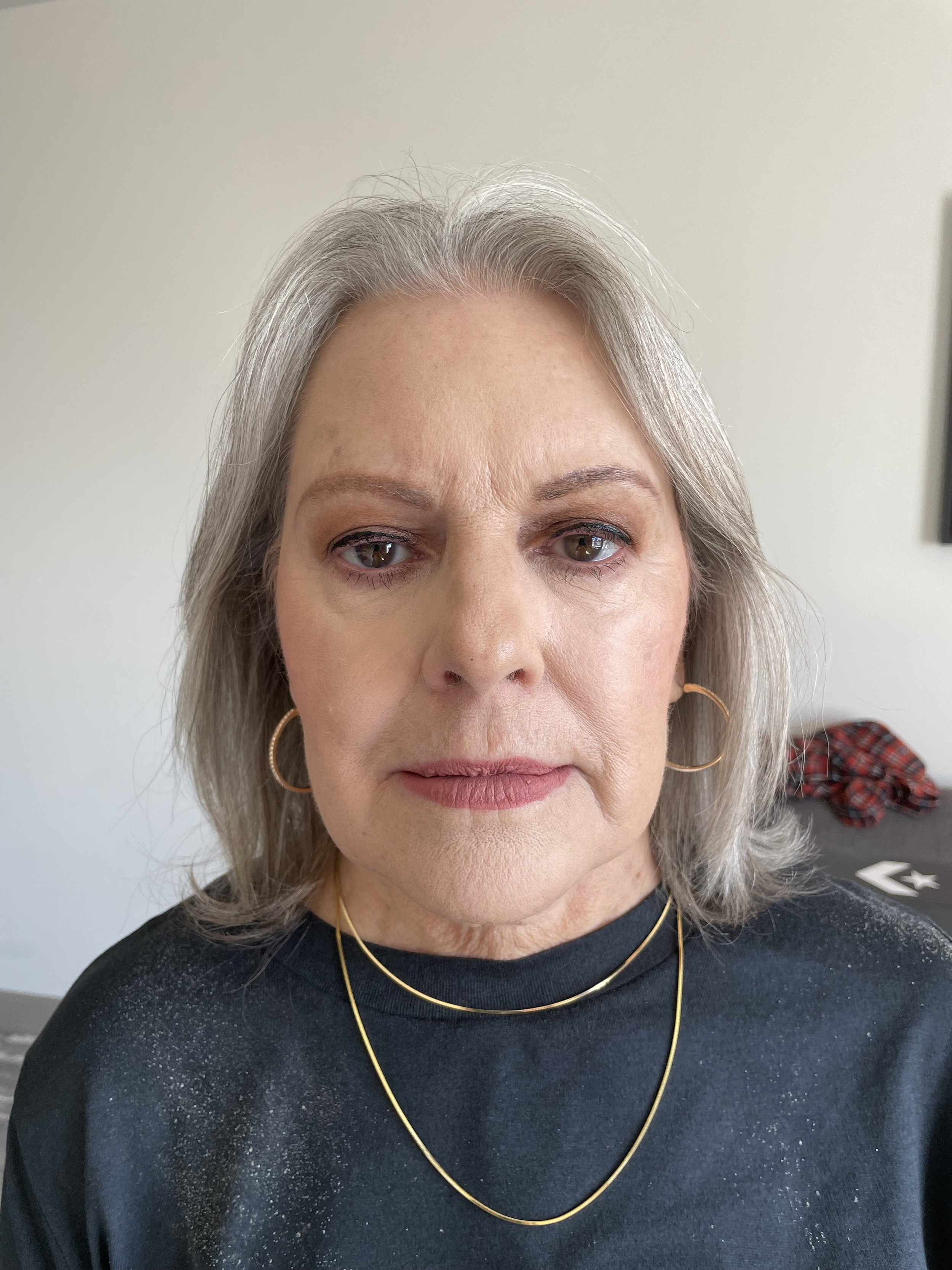 Close-up of an older woman with gray hair, wearing hoop earrings and gold necklaces, looking directly at the camera.