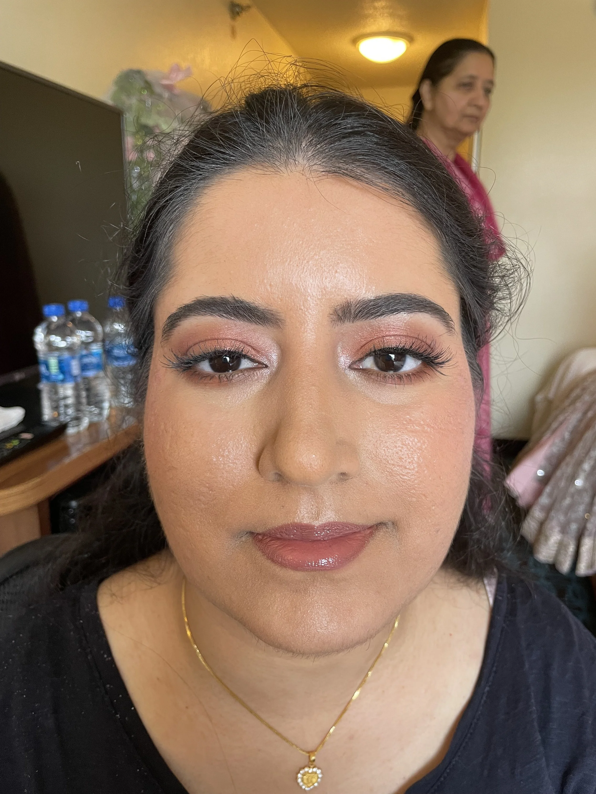 Close-up of a woman with makeup, wearing a gold necklace with a heart-shaped pendant, sitting indoors with another woman in the background near a TV and water bottles on a table.