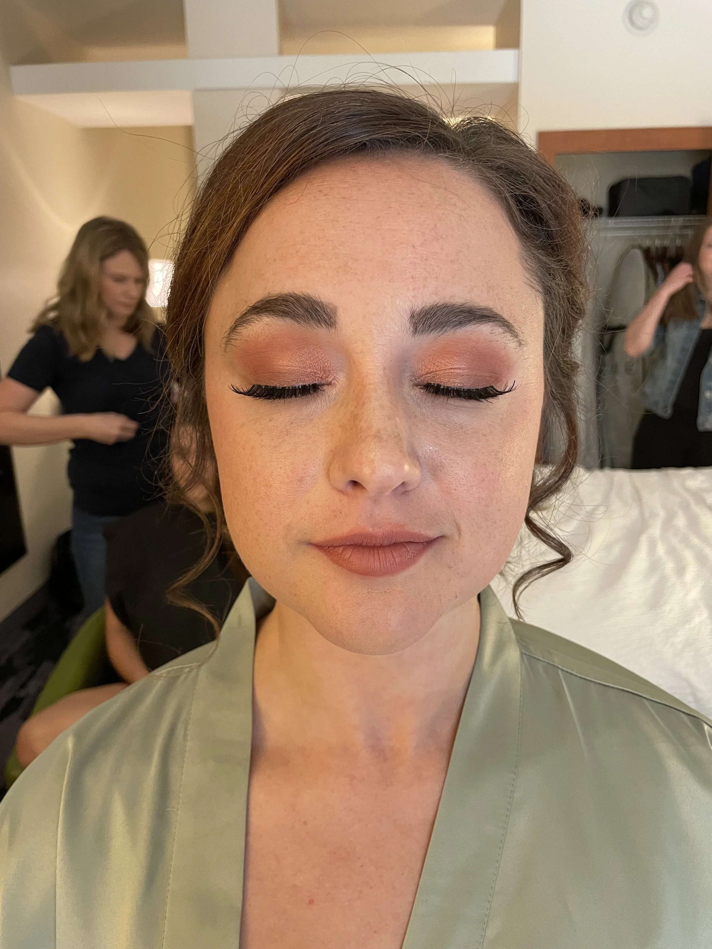 Close-up of a woman with her eyes closed, wearing makeup, and a satin robe, in a room during getting ready.