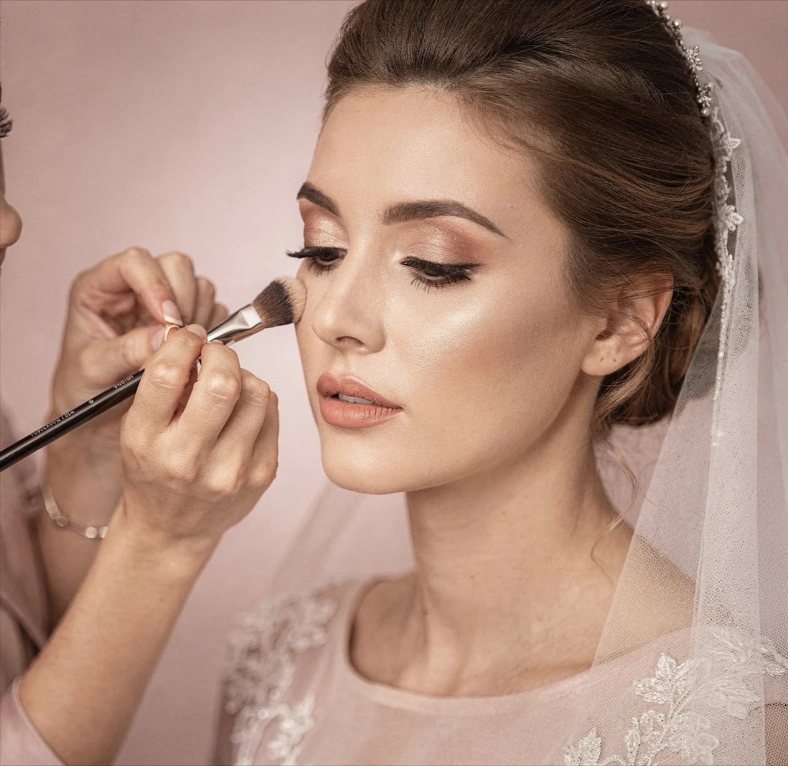 Close-up of a bride with brown hair and light makeup, having her makeup applied by a makeup artist, who is using a brush on her cheek. The bride is wearing a lace wedding dress and a veil.