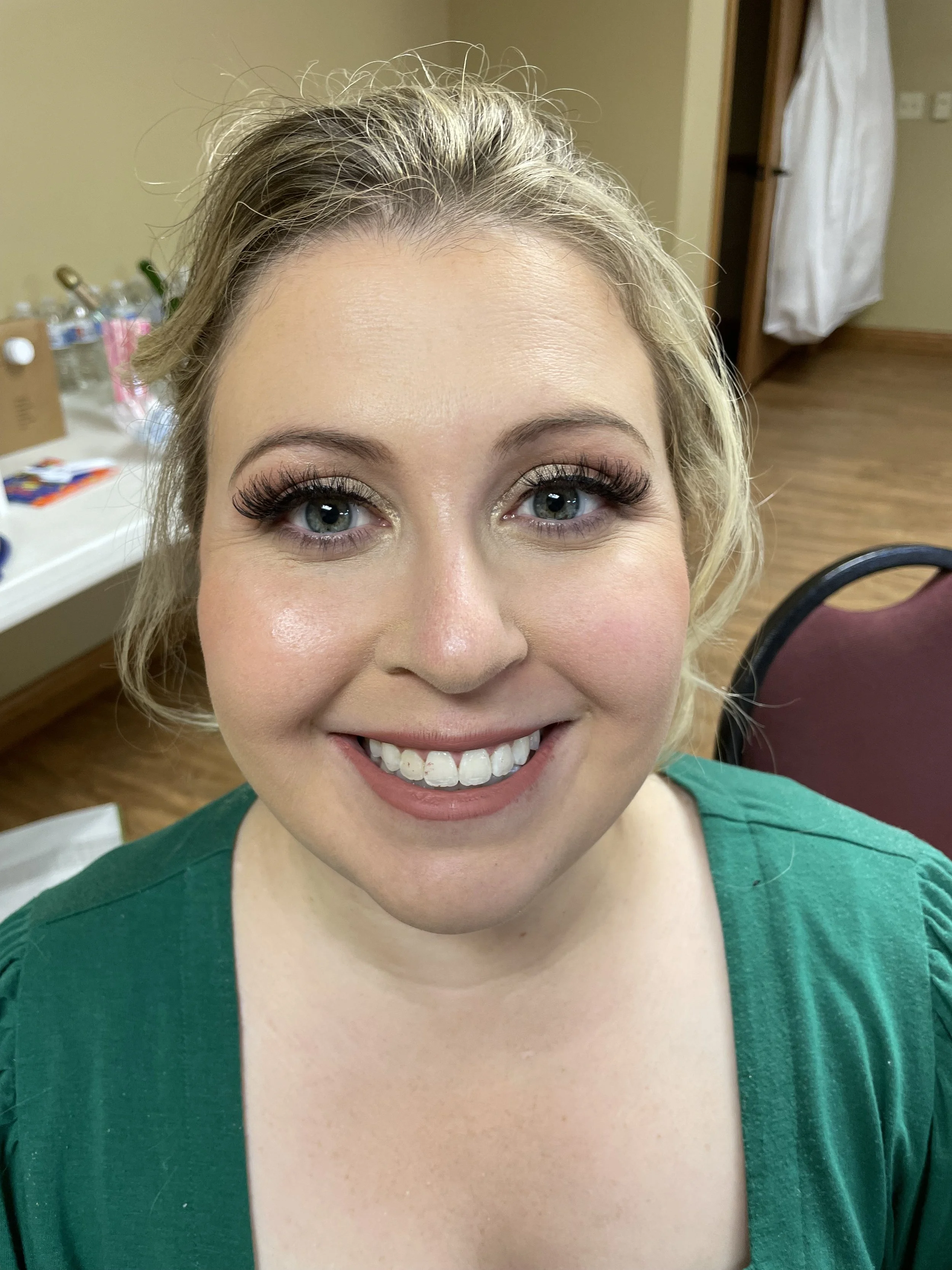 A smiling woman with blonde hair, wearing makeup, and a green top, in  a bridal getting-ready room.