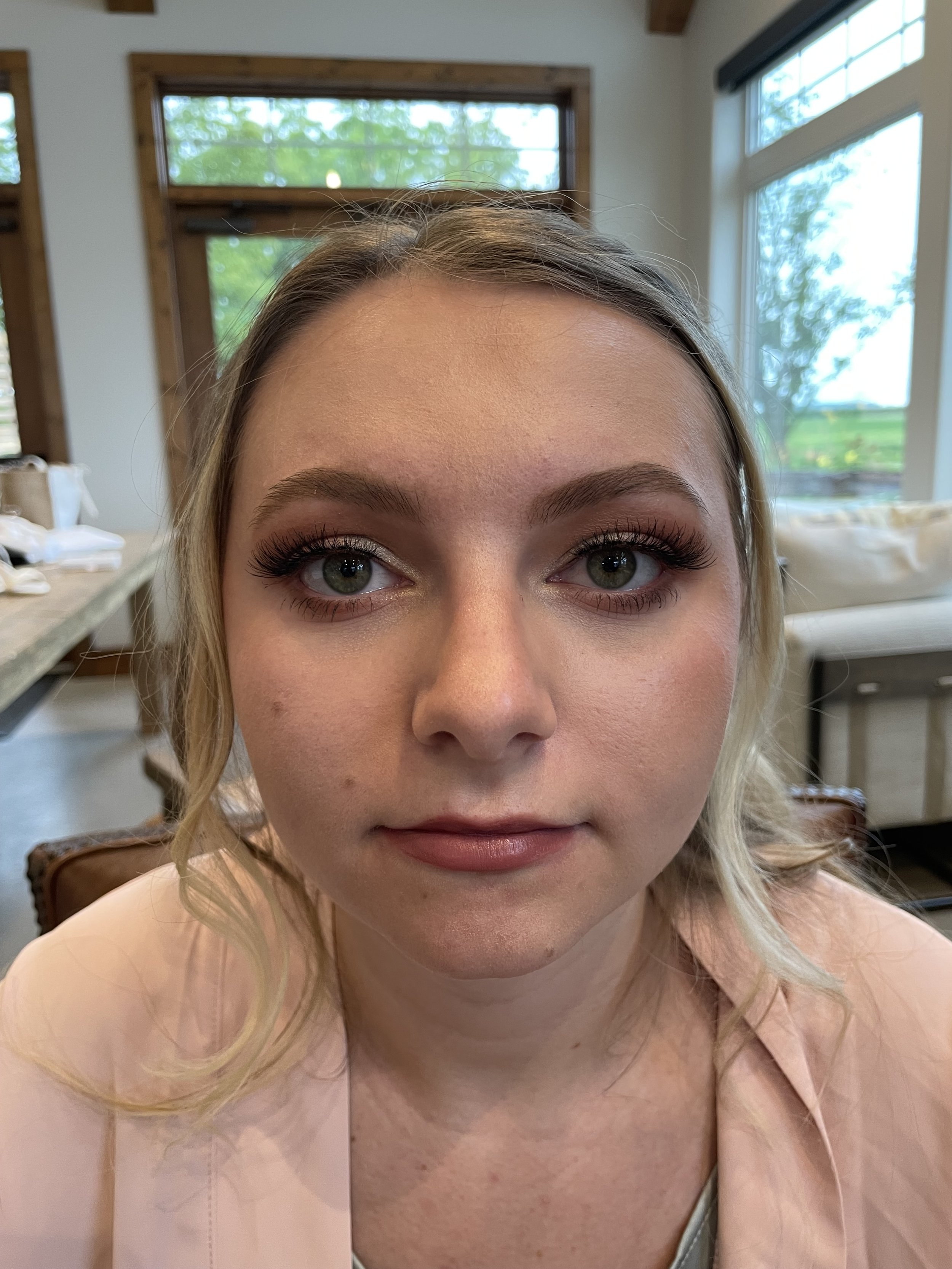 Close-up of a woman with light skin, blonde hair, and makeup, sitting indoors with large windows showing a green, outdoor landscape.