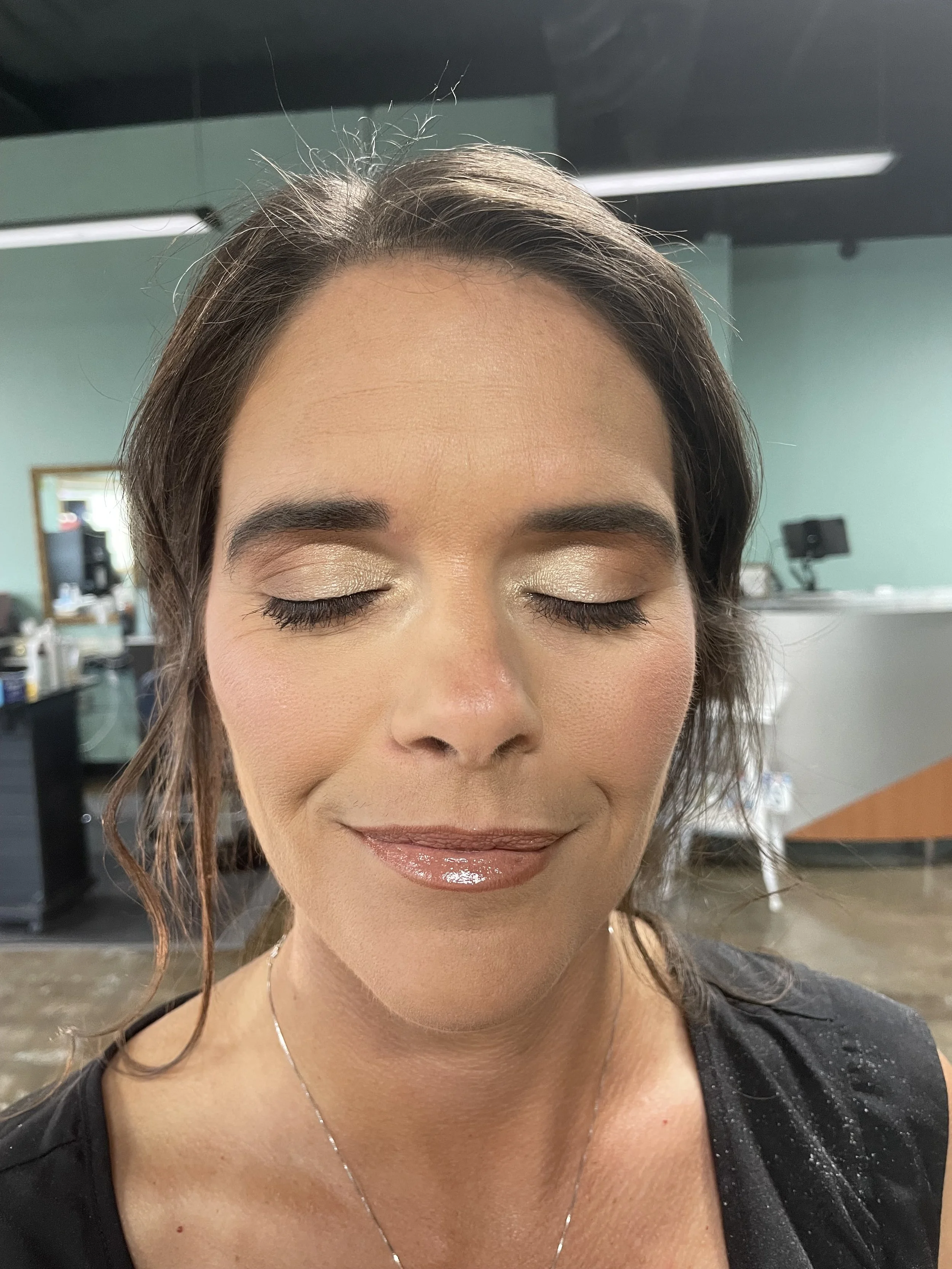 Close-up of a woman with closed eyes, wearing makeup with gold eyeshadow, glossy lips, and a black top, in a room with salon or studio background.