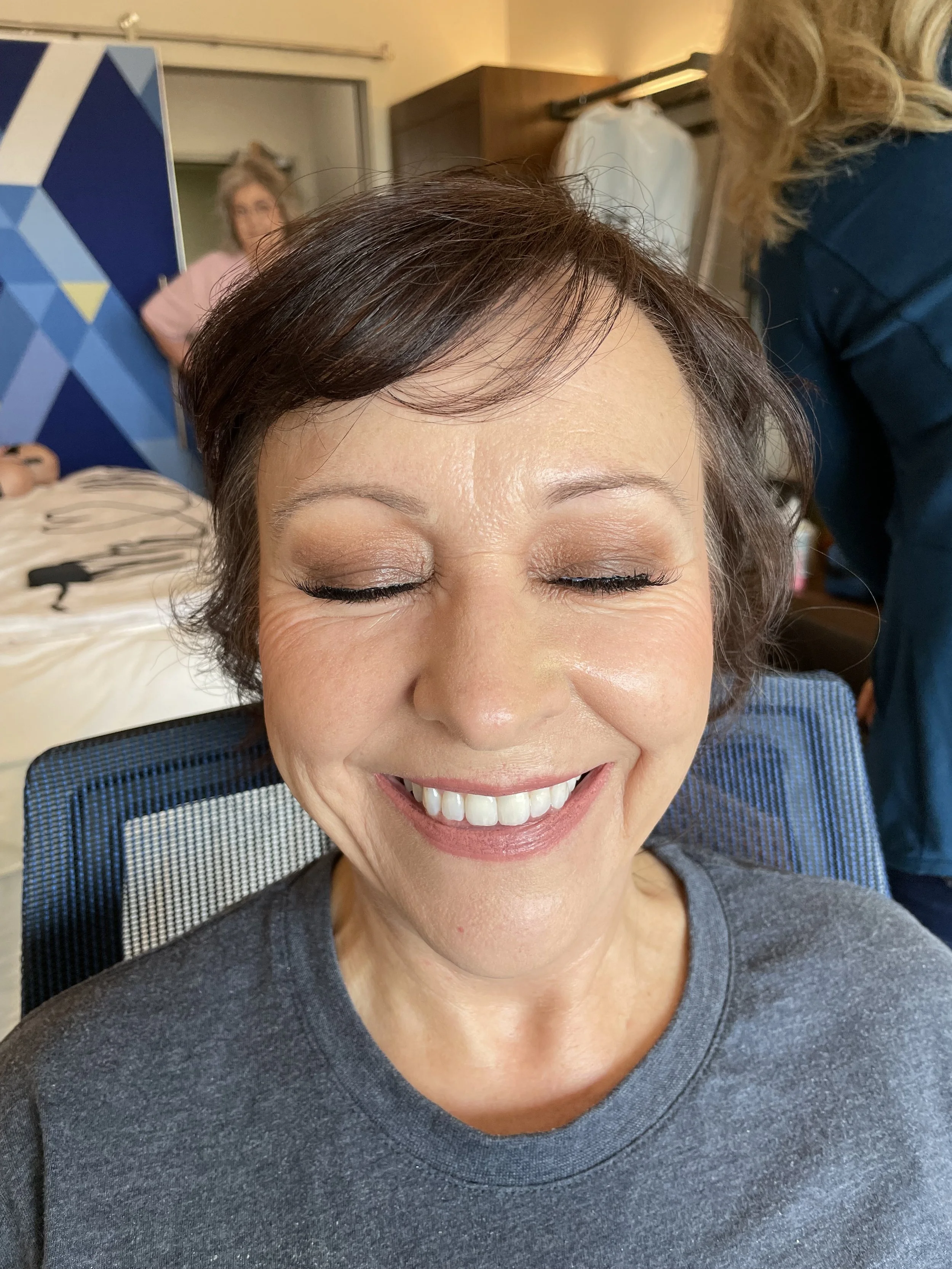 A mother of the bride/groom with short brown hair and closed eyes, smiling at the camera showing off her makeup.