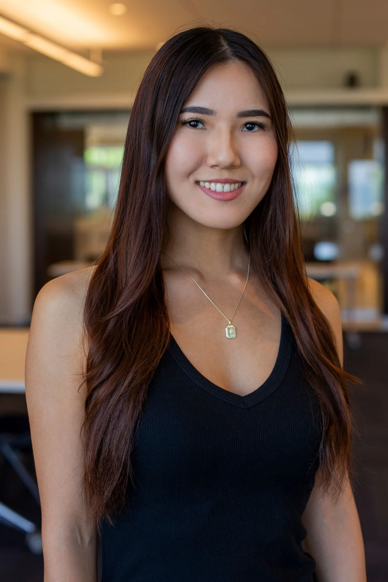 A young woman with long brunette hair smiling, wearing a black sleeveless top and a necklace with a pendant, standing indoors with a blurred background.