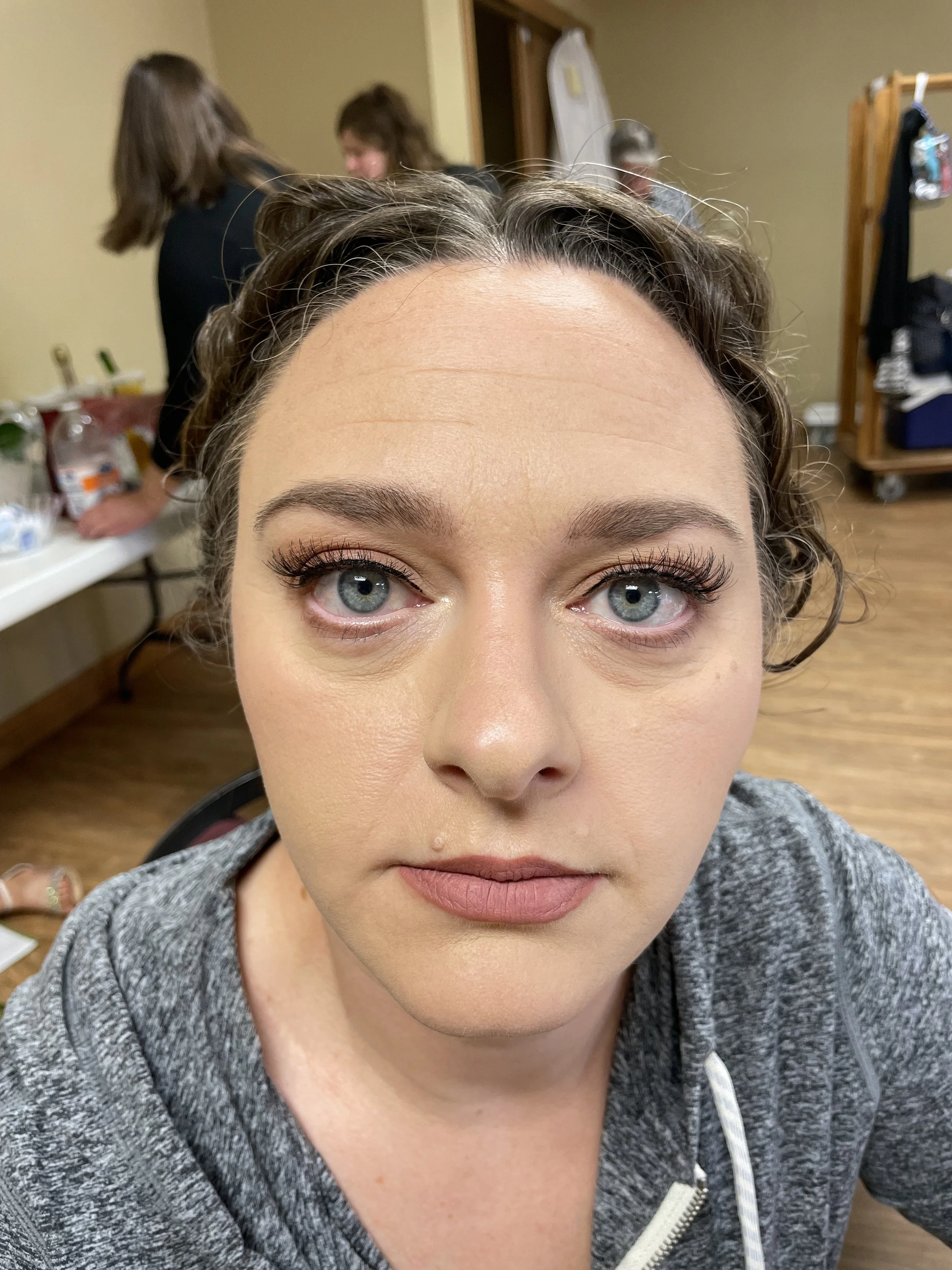 Close-up of a woman with blue eyes, makeup, and curly brown hair in a dressing room.